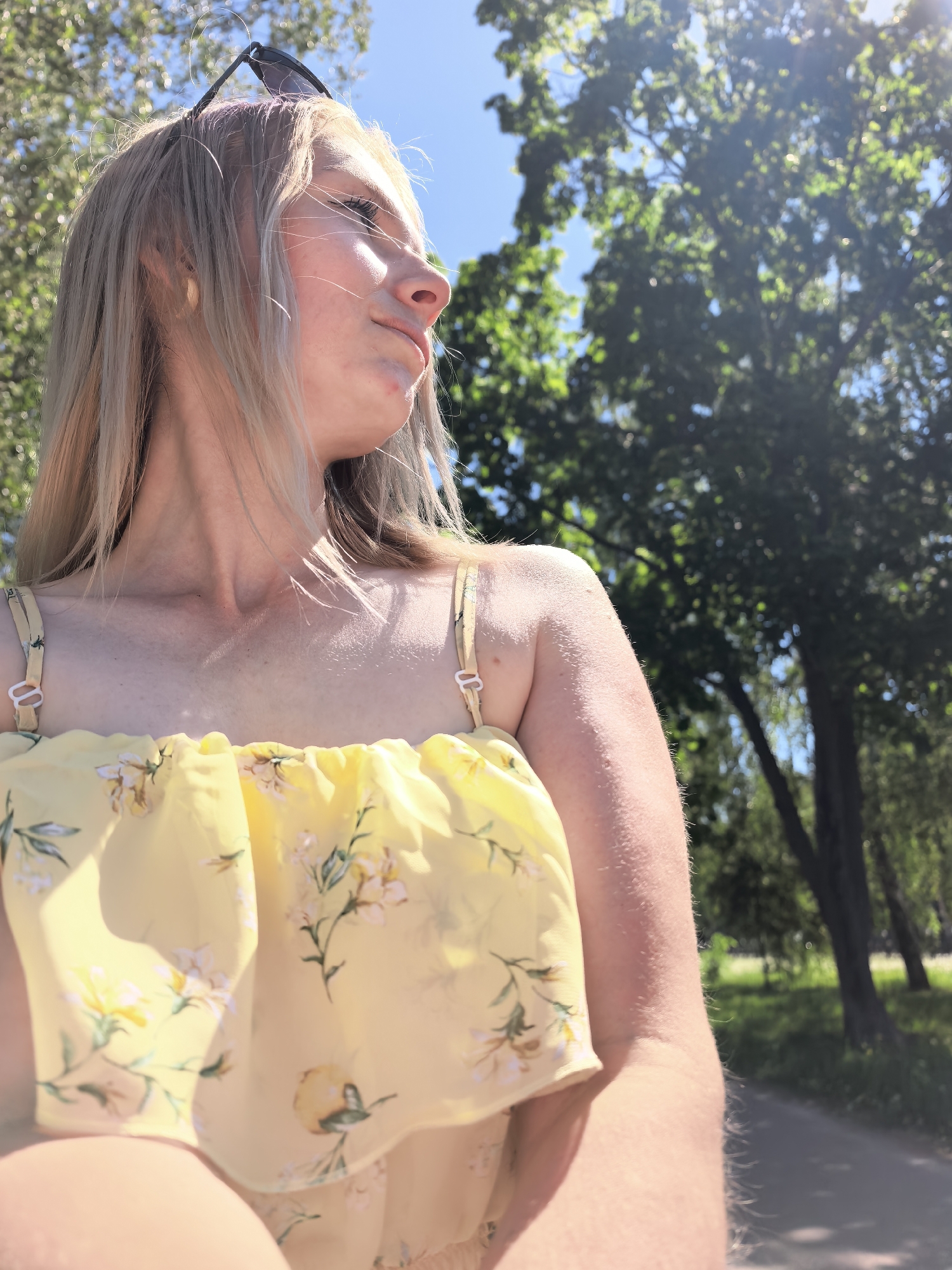 A young woman with blonde hair and sunglasses perched on her head is looking to the right of the frame. She wears a yellow floral patterned top with thin straps. The background shows green trees and a clear blue sky, suggestive of a sunny day in a park. The location is likely a park area within Sterlitamak, Russia. A gray road is visible at the edge of the trees.