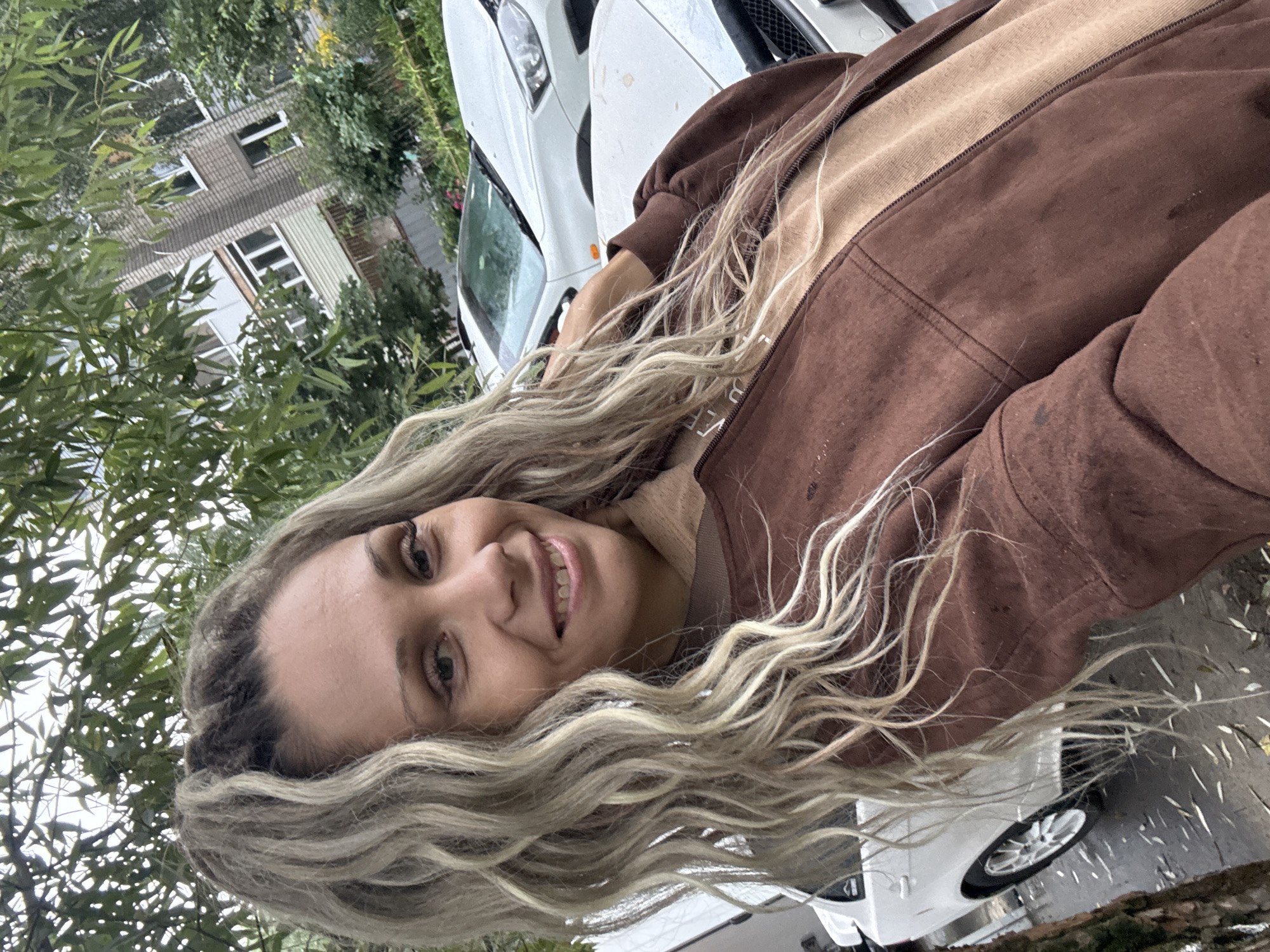 A blonde woman with long, wavy hair smiles at the camera. She is wearing a brown jacket over a tan shirt. In the background, a white car is parked in front of a brick building with trees and foliage. The scene appears to be outdoors in a residential area.