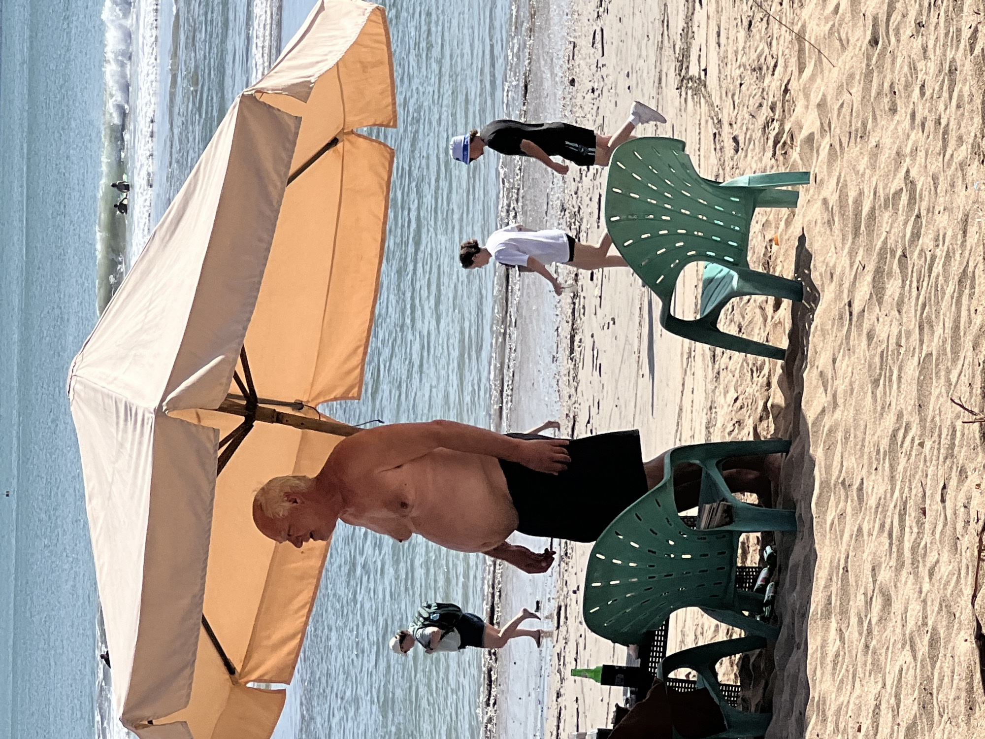 In Kuta, Indonesia, a man stands shirtless under a large, beige beach umbrella on a sandy shore. He is a white male with a prominent belly, wearing black shorts. To his left, two green plastic chairs and a black table with a green bottle are positioned on the sand. In the background, the ocean with gentle waves is visible, and several people are walking along the shoreline. A woman with a backpack walks towards the left, and further out, a couple walks near the water's edge, one wearing a white t-shirt and the other a black t-shirt and a blue bucket hat. Two surfers are visible in the water further out. The sand is light brown and textured with ripples.