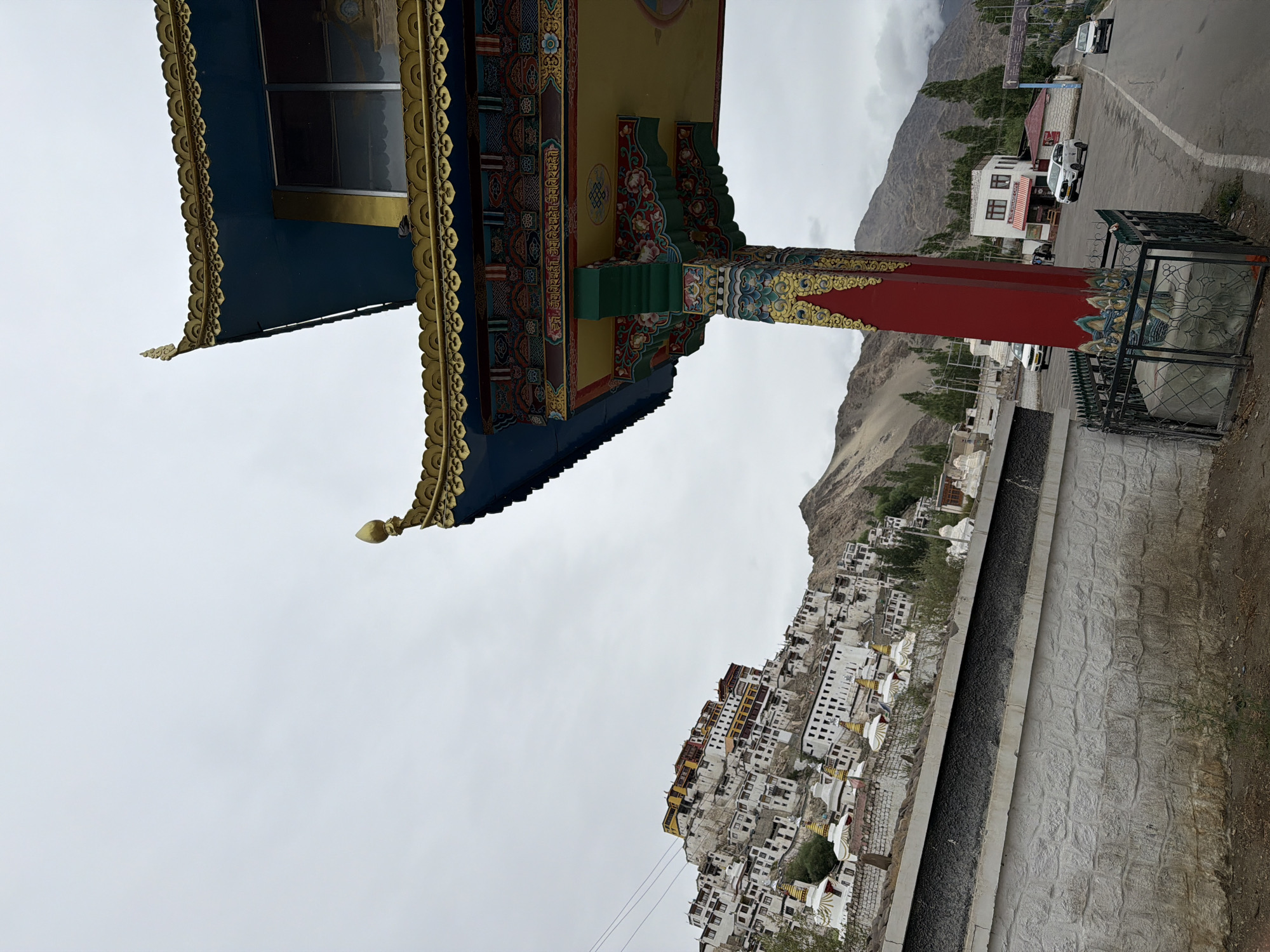 In the city of Thiksey, India, a detailed, matter-of-fact description of the scene reveals a multi-tiered Buddhist monastery complex perched on a hillside under a cloudy sky. The monastery's whitewashed buildings, adorned with yellow and red roofs, cascade down the rocky incline, interspersed with stupas. In the foreground, a vibrant, ornate gateway structure with a blue roof and intricate gold and multi-colored detailing stands prominently, supported by a red pillar. A paved road runs alongside the gateway, with several white cars parked and driving on it. A low stone wall with a textured surface lines the foreground, separating the road from the base of the monastery. The natural environment includes barren, mountainous terrain surrounding the settlement and scattered green trees. The overall impression is one of a significant cultural and religious site within a rugged landscape.