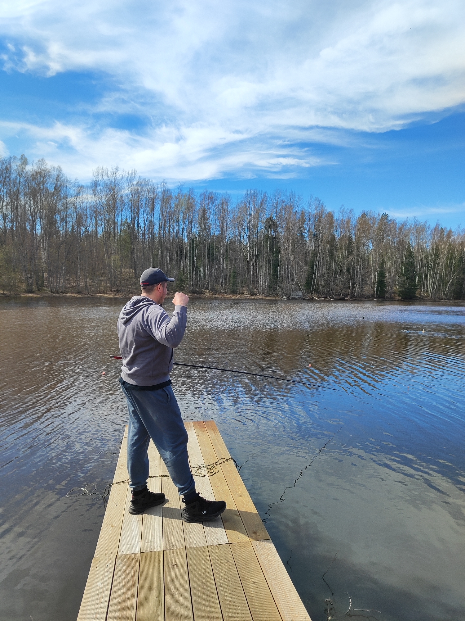 A person stands on a wooden dock, fishing in a lake. The person is wearing a gray hooded sweatshirt, gray sweatpants, black shoes, and a dark cap. They are holding a fishing rod. The lake is surrounded by trees, some of which are bare. The sky is blue with white clouds. The location appears to be a natural area, possibly a park or recreation area. The city is Unknown, Unknown.
