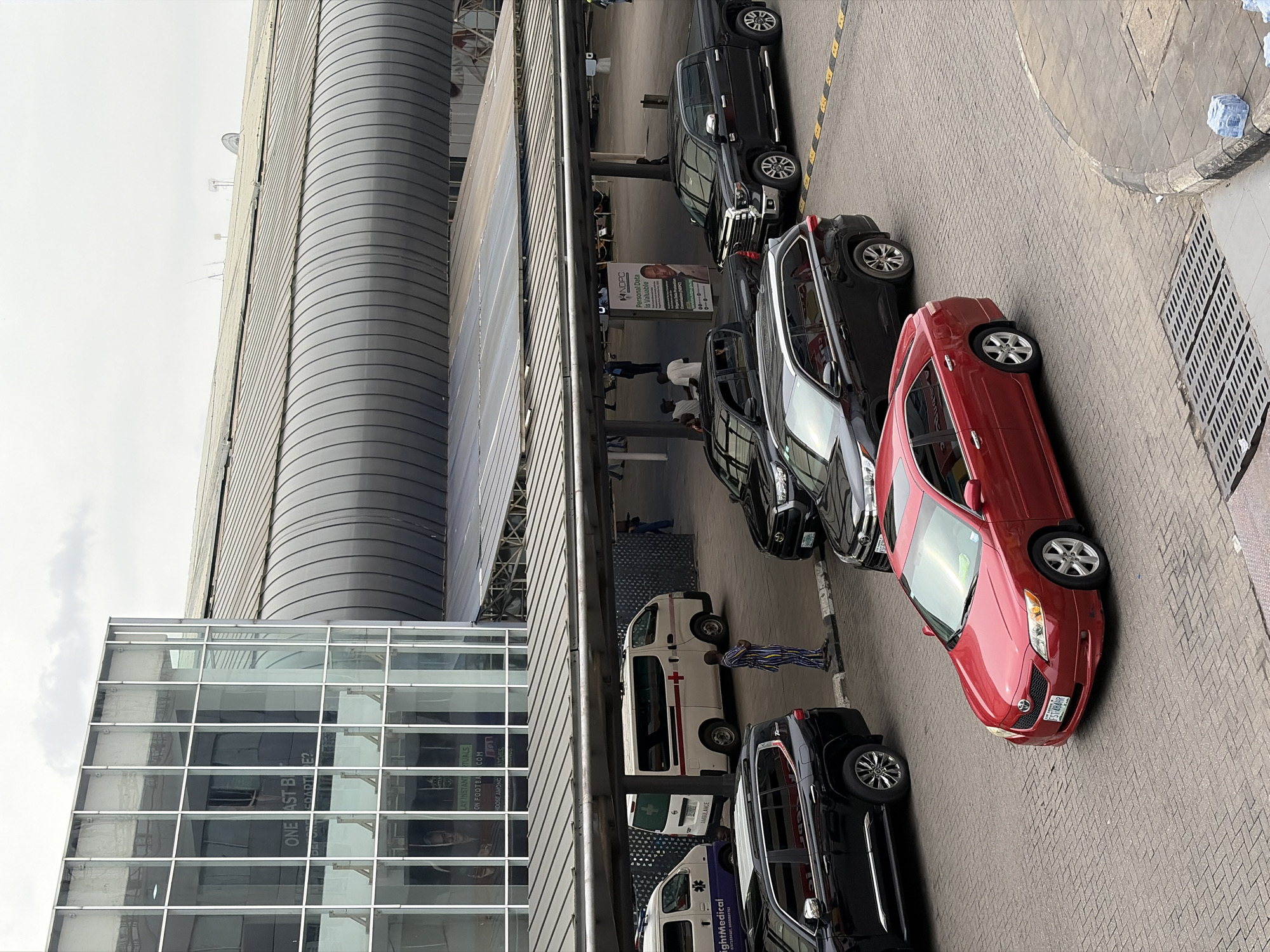 The image depicts an urban scene in Ikeja, Nigeria, featuring a modern building with glass windows adjacent to a curved, metal-clad structure. Several vehicles are parked along the roadside, including black SUVs, a red sedan, and at least two ambulances, one marked "RightMedical." A person wearing striped clothing stands near one of the ambulances. Signage with the acronym "NDPC" is visible. The ground is paved with interlocking stones, and a drainage grate is present on the right side. The sky is overcast.