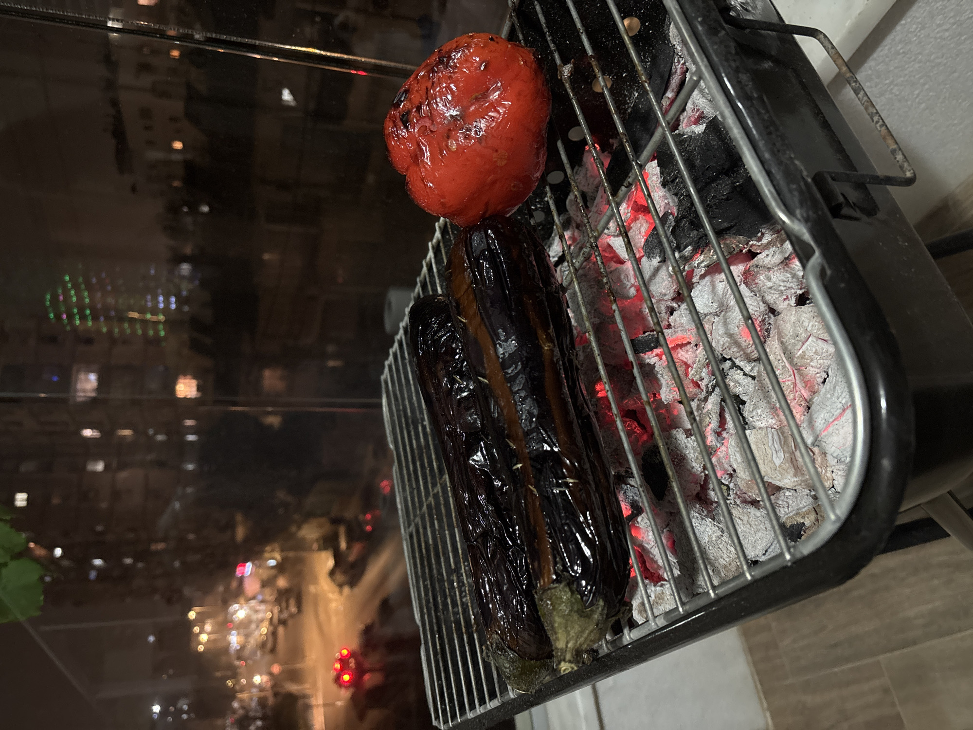 A top-down, close-up shot shows a portable charcoal grill with a red bell pepper and two eggplants being grilled. The charcoal is white and red hot. The background shows the city of Bat Yam, Israel, at night through a window. City lights and traffic are visible. The grill is on a tiled surface.