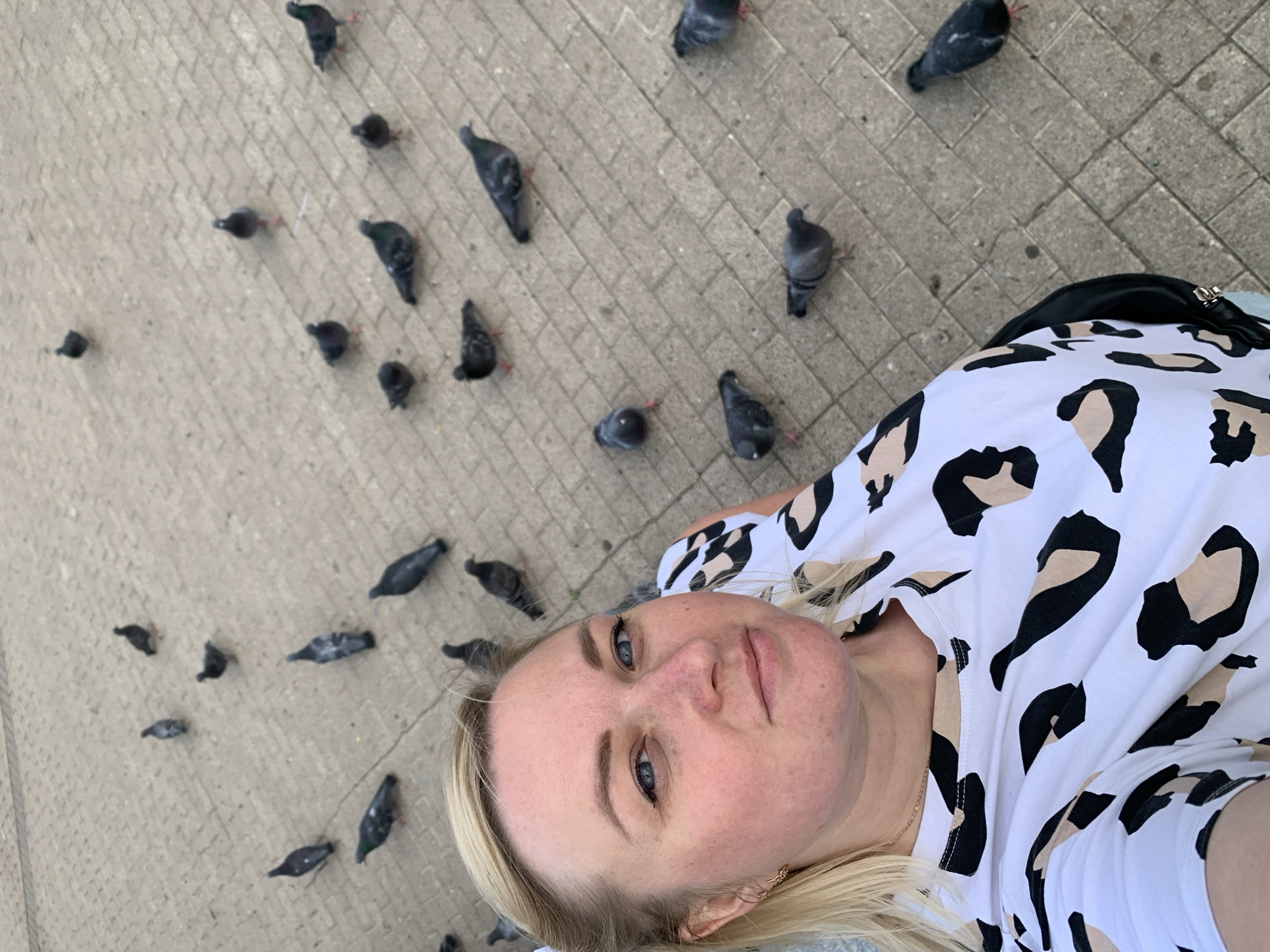 The image captures a selfie-style shot of a blonde woman with blue eyes, wearing a white shirt with black and tan animal print, lying on a brick sidewalk. The location is populated with numerous pigeons, scattered across the paving stones. The background suggests an urban environment, likely in a public square or pedestrian area. No specific details confirm the location as Chaikovsky, Russia.