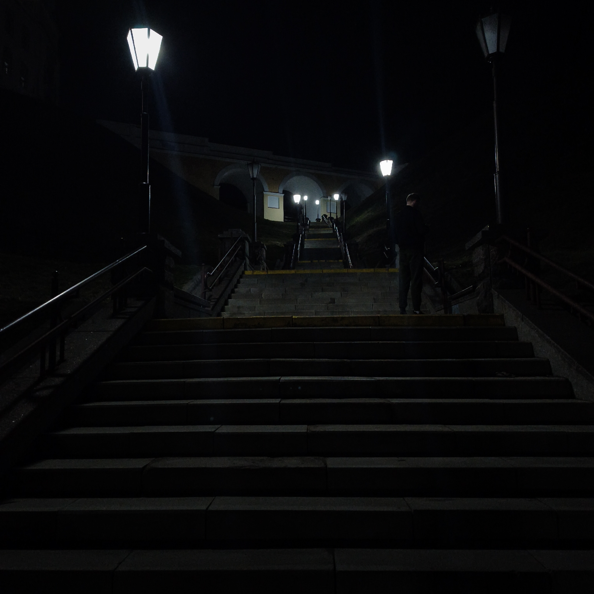 The image depicts a nighttime scene of a grand outdoor staircase in Hrodna, Belarus, specifically the ascent leading to the Old Castle (Stary Zamek). The multi-flight staircase, constructed of dark stone or concrete, dominates the center of the frame, ascending towards a prominent, multi-arched, yellowish-beige architectural structure at the top.
One male individual, viewed from behind, stands on a landing approximately midway up the visible staircase. He wears dark trousers and a dark upper garment, possibly a jacket or shirt. His posture suggests he is paused or in the process of ascending.
The scene is illuminated by several streetlights. Two brightly lit, white, hexagonal-lantern streetlights are prominent on either side of the lower-to-mid sections of the stairs. Additional, smaller streetlights are visible further up the staircase, casting light towards the arched structure. Dark metal handrails run along the sides of the stair sections. Some steps feature yellow visibility strips along their edges. The environment is otherwise dark, with indistinct dark masses suggesting sloped terrain flanking the staircase.