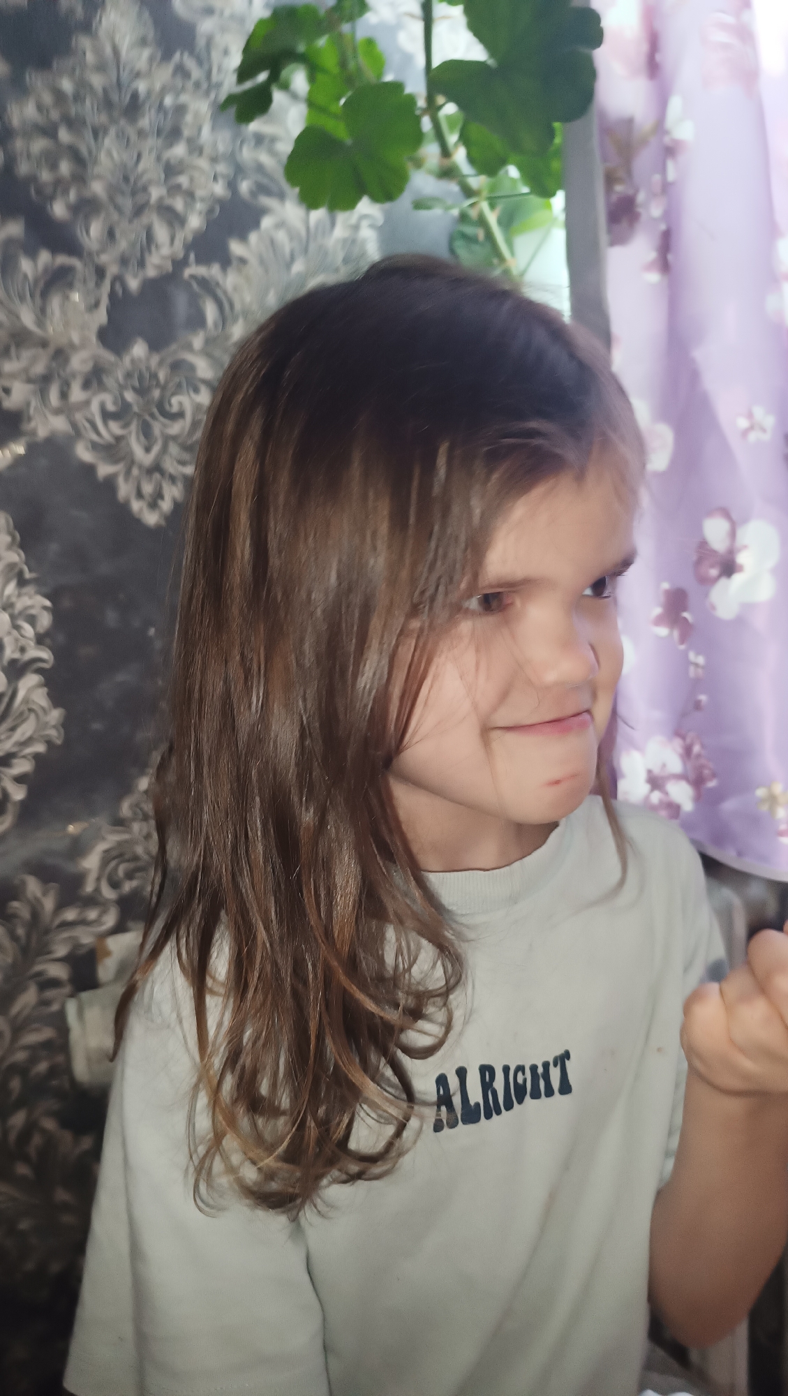 A young girl with long brown hair and hazel eyes is looking to her right with a slight smile and her mouth slightly open. She is wearing a light-colored t-shirt with the word "ALRIGHT" printed in dark blue letters across the chest. Her right hand is partially visible in the lower right corner, appearing to be held up near her face. The background features a patterned wallpaper on the left and a sheer purple curtain with floral designs on the right. A potted plant with green leaves is visible behind the girl, near the top center of the image. The scene is indoors, with soft lighting that appears to be coming from the right. There are no discernible landmarks or environmental cues that would indicate a specific location in the city of Karsha, Russia.