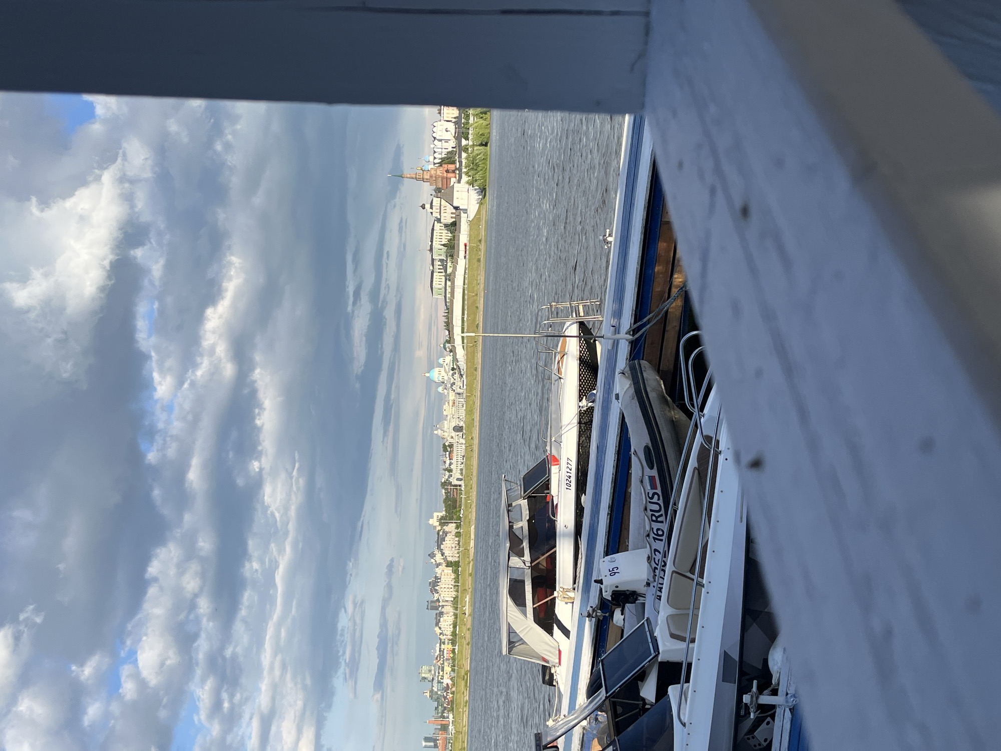 The image depicts a view of Kazan, Russia, from a boat or a dock. In the foreground, a white wooden structure partially obscures the view. Several boats are docked next to the viewer, including one with the Russian flag and "16 RUS" visible on its hull. In the background, across the water, lies the skyline of Kazan, including the Kazan Kremlin with its distinctive towers and blue-domed cathedral. The sky is filled with gray and white clouds.