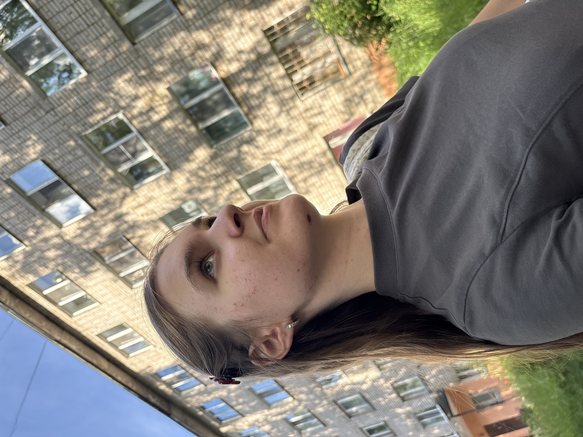 A close-up, vertical shot features the upper body and face of a young person outdoors. The individual is looking upwards and to the left, with a neutral expression. Their face is partially illuminated by sunlight, revealing freckles and some blemishes. They have long, brown hair, some of which is pulled back with a dark red clip. A small, silver hoop earring is visible in their left ear. They are wearing a dark grey hooded sweatshirt.
The background is out of focus, but appears to be a building with multiple windows and brickwork, possibly a residential building. Sunlight casts shadows from an unseen object, likely trees, onto the building's facade. To the right, there is greenery, suggesting a park or garden area. The sky is visible in the lower portion of the image, appearing blue and clear. The orientation of the image suggests it was taken from a low angle, looking up. The lighting and shadows indicate it is daytime, likely sunny.
The overall scene suggests a casual outdoor setting, possibly in a city or town in Chaikovsky, Russia, given the context provided. No text is visible in the image.