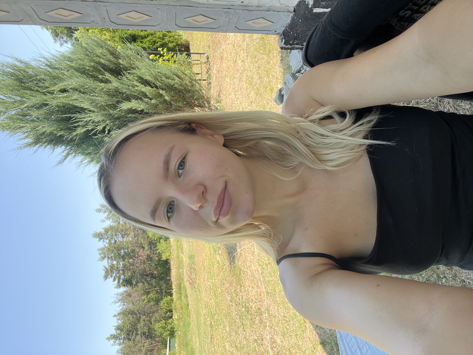 A young woman with blonde hair and blue eyes is taking a selfie outdoors. She is wearing a black tank top. She is sitting in what appears to be a rustic outdoor setting, with a field and trees in the background. A tall evergreen tree is to her right, and a portion of a weathered structure with diamond-shaped patterns is visible on the far right. The sky is clear and blue.