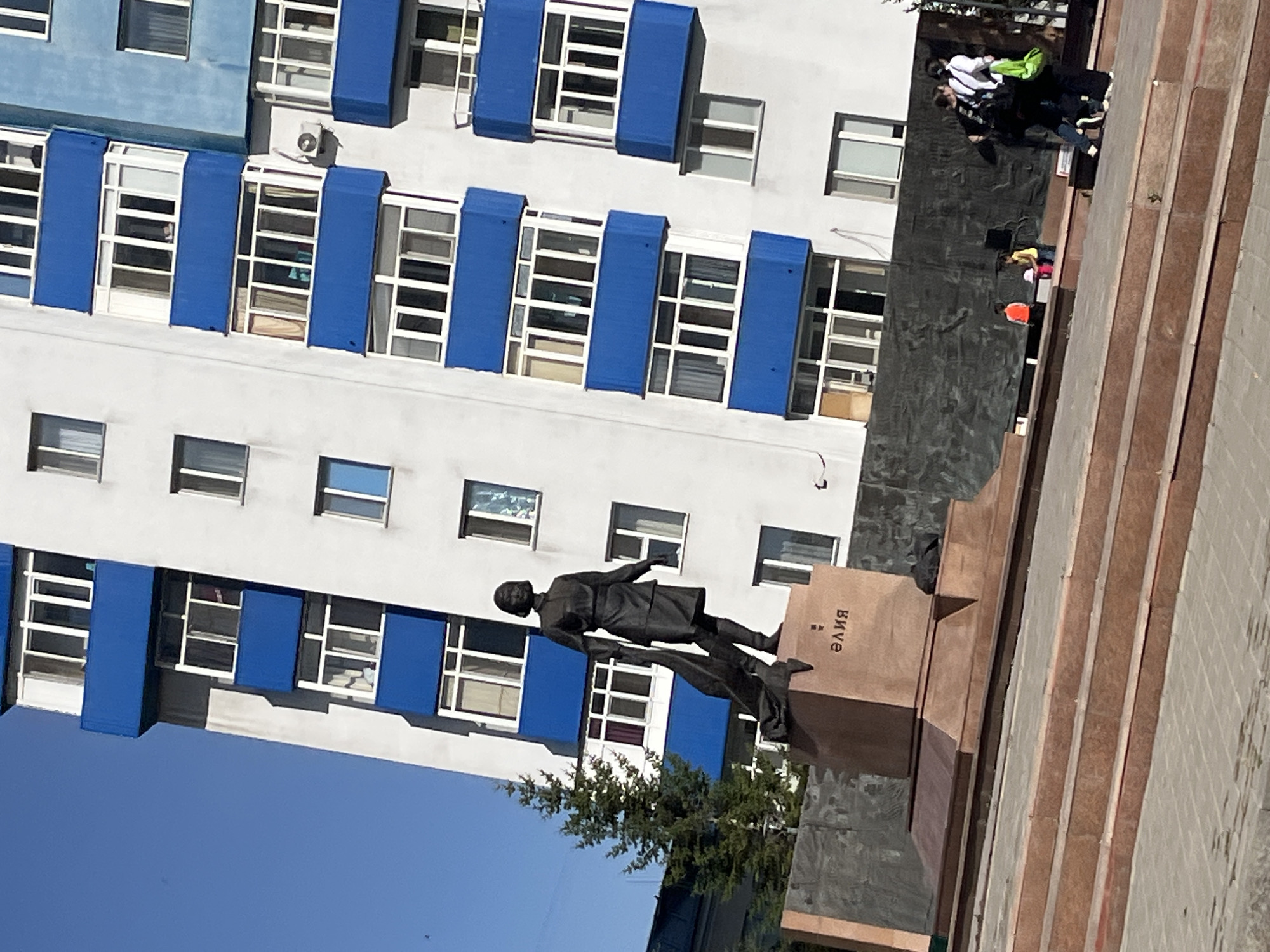 The image captures a monument featuring a statue of a woman in a dark outfit, standing on a pedestal. The pedestal has Cyrillic lettering inscribed on its side, reading "элия". Behind the monument, a modern apartment building with white and blue accents dominates the skyline, characterized by numerous windows and balconies. A tall evergreen tree stands to the left of the monument. In the foreground, stone steps lead up to the monument area. A group of people are visible near the monument, some walking and others standing, including individuals carrying bags. The scene appears to be an outdoor public space, likely a square or plaza, in the city of Astana, Kazakhstan.