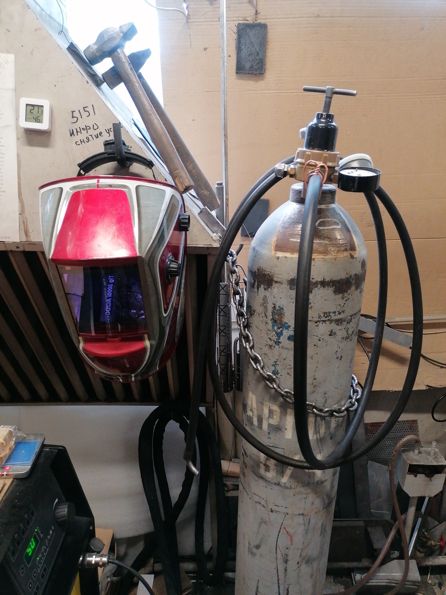The image shows a cluttered workshop setting. A gray gas cylinder secured with a chain is prominently displayed, connected to a black hose and a regulator with a gauge. Adjacent to the cylinder, a welding helmet hangs, revealing a screen with text in the inside. Two hammers are mounted on a shelf above the helmet. A digital thermometer is affixed to the wall with "21.3" showing, and "46" for humidity. Graffiti-like markings appear next to the thermometer, featuring numbers and lettering. In the lower-left corner, part of a welding machine is visible. The environment is characterized by a disorganised array of equipment and materials.
