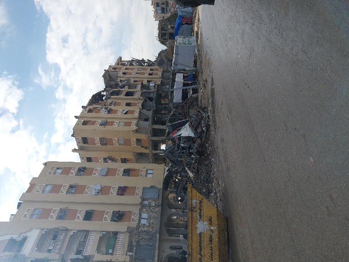 This image depicts widespread structural damage in Khan Yunis, Palestinian Territory. In the midground, multiple multi-story buildings, likely residential, display extensive destruction. Their facades, originally pale yellow with decorative trim, are now scarred with shattered windows, damaged balconies, and visible structural compromises, particularly on lower floors where sections have collapsed. Large piles of concrete rubble, twisted metal, and debris are accumulated at the base of these buildings and extend into the foreground, partially obscuring the lower levels.
On the left, a large yellow industrial dumpster with Arabic script is present on the edge of a wet, asphalt road that dominates the foreground. To the right, beyond the primary rubble pile, several makeshift shelters or market stalls constructed from tarpaulins (blue and white) are visible. A person's legs and lower torso, clad in dark clothing, are partially visible on the extreme right edge of the frame, facing away from the viewer. The sky above is partly cloudy with patches of blue visible. The overall scene indicates significant devastation.