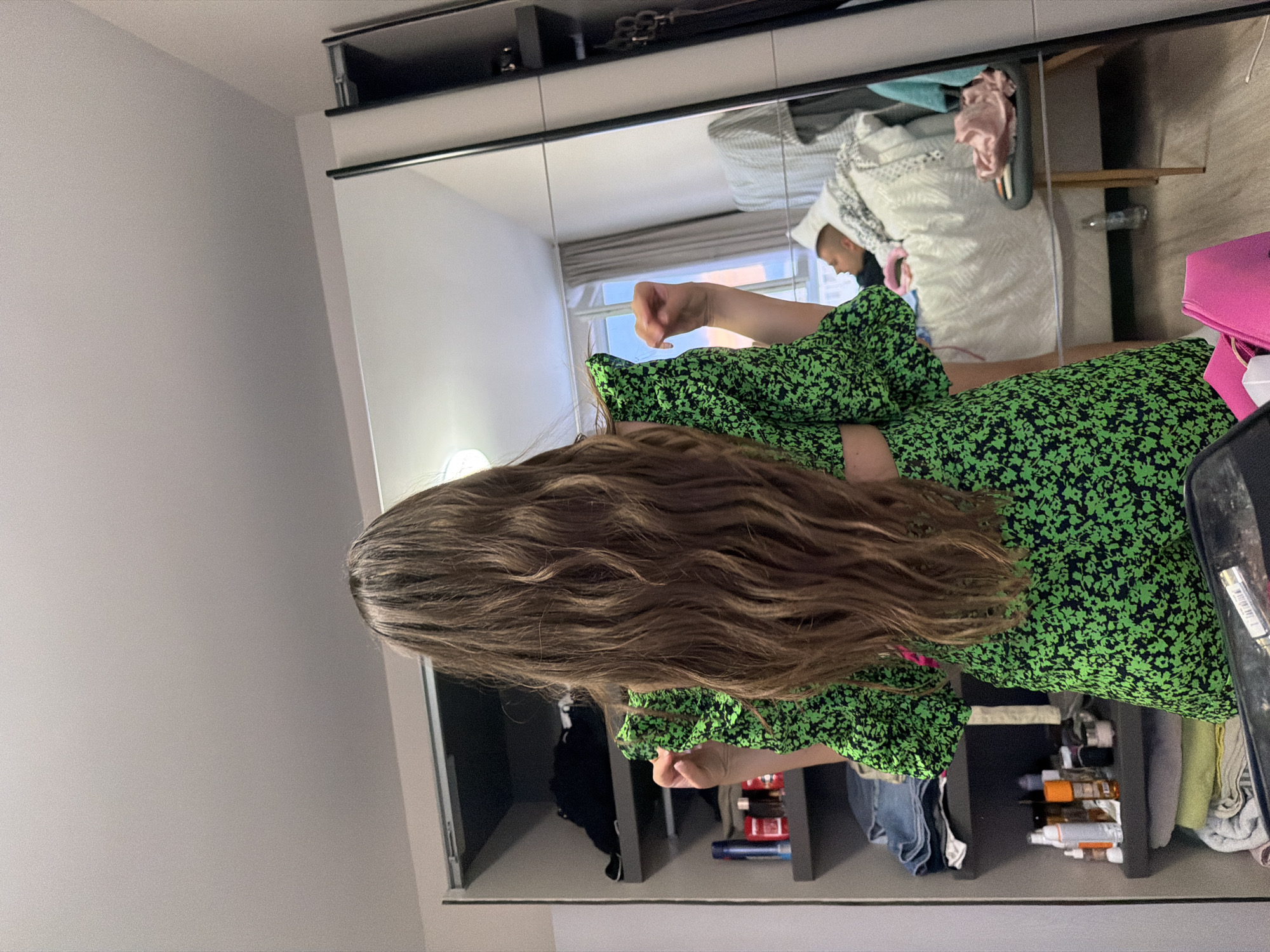 The image depicts an indoor scene, likely a bedroom or dressing area, in the city of Plovdiv, Bulgaria. In the foreground, a person with long, wavy brown hair, seen from behind, is standing, facing a large mirror on the right side of the frame. The person is wearing a short-sleeved green dress with a black floral pattern and appears to be in the process of adjusting it or posing, with both hands slightly raised and bent at the elbows.
To the left of the person, an open shelving unit or wardrobe with dark grey compartments is visible. The shelves contain various items: the top visible shelf has folded dark clothing; the second shelf down has folded denim jeans; the third shelf displays an assortment of personal care products including a blue aerosol can, a red bottle, and several smaller bottles and jars; the bottom visible shelf holds stacks of light-colored towels or linens.
The large mirror on the right reflects a significant portion of the room behind the viewer. In the reflection, a male individual with short hair is lying on a bed, covered with white bedding, appearing to be resting or sleeping. Behind the bed, a window with light-colored curtains (beige/grey) is visible, with glimpses of an exterior view. A small, dark-upholstered stool with light wooden legs is positioned next to the bed, holding a folded pink garment. A clear plastic water bottle is on the floor beside the stool. A part of a pink bag belonging to the main subject is also reflected in the lower right corner of the mirror.
The floor is a light-colored wood or laminate, and the ceiling is plain and white. The lighting appears to be a mix of natural light from the window and ambient indoor light.