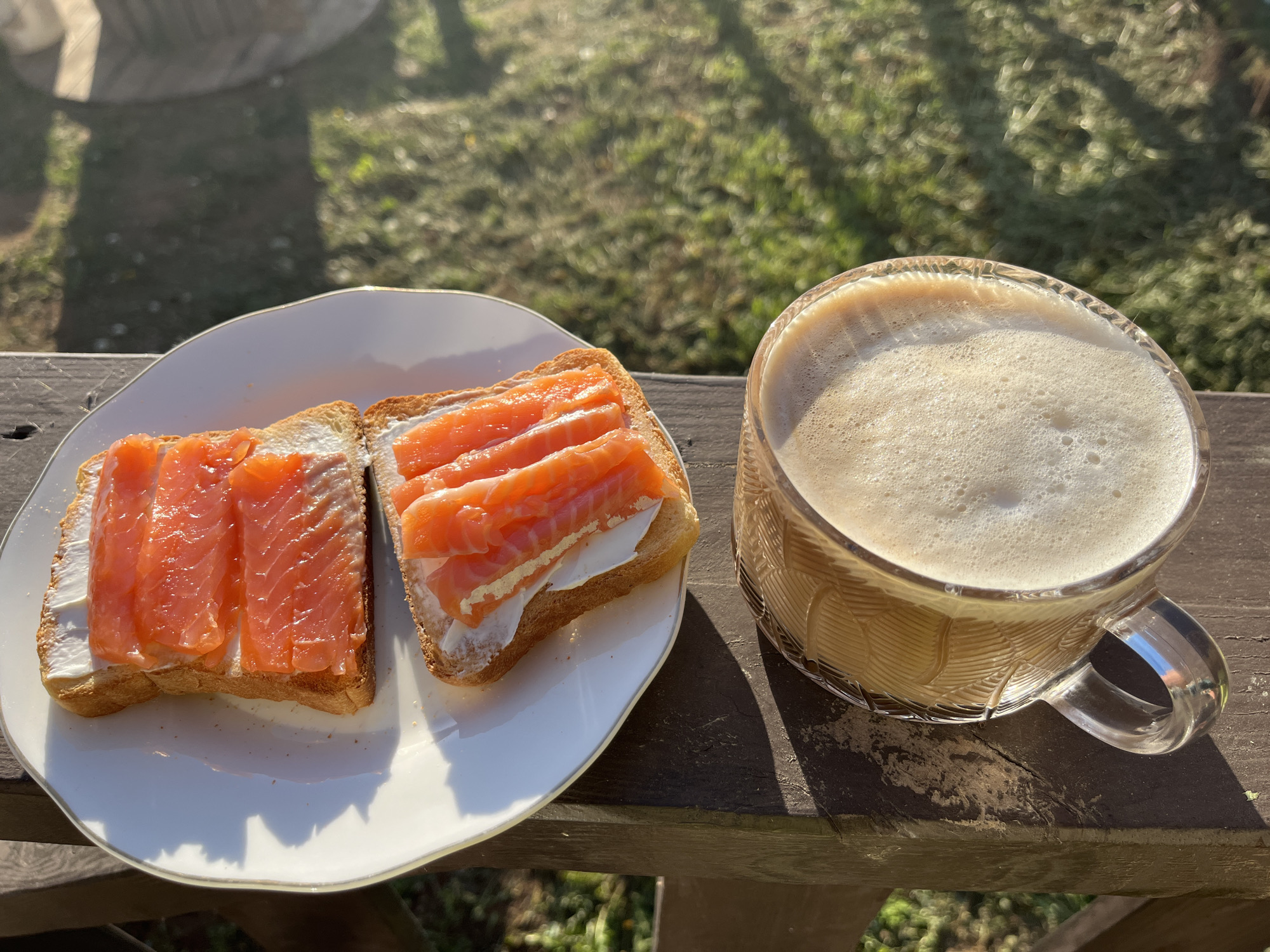 A white plate with two pieces of toast topped with cream cheese and sliced salmon is positioned next to a glass mug filled with a foamy beverage, likely coffee or latte. The scene is set outdoors on a wooden railing or deck, with blurred greenery visible in the background. The lighting suggests it is daytime. The image does not depict any people or actions. It appears to be a still life arrangement of food and drink. There is no evidence to indicate the city of Chaikovsky, Russia.