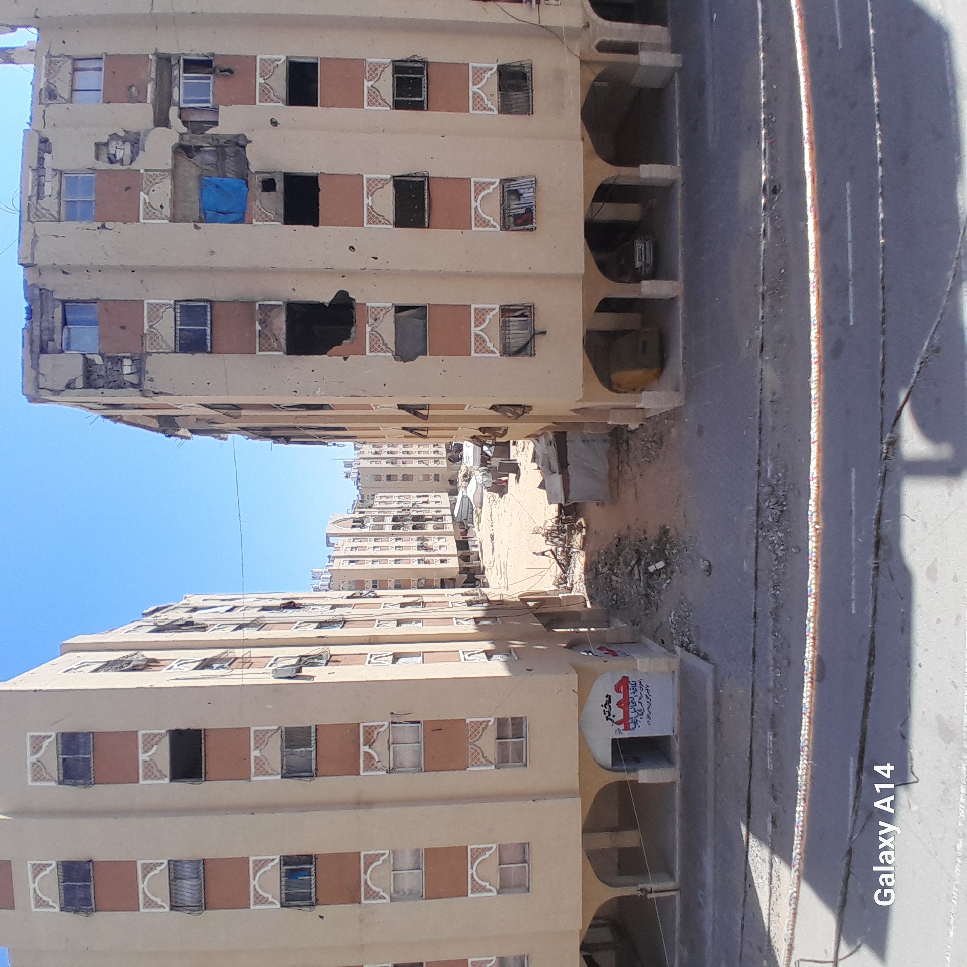 The image depicts a street scene in what appears to be a Middle Eastern city, characterized by tall, tan-colored apartment buildings on either side. The buildings show signs of damage, including missing window panes and structural degradation, indicative of conflict or neglect. The street between the buildings is littered with debris, reinforcing the impression of a distressed environment. The sky is clear and blue. The photograph includes a "Galaxy A14" watermark, indicating the device used to capture the image.
