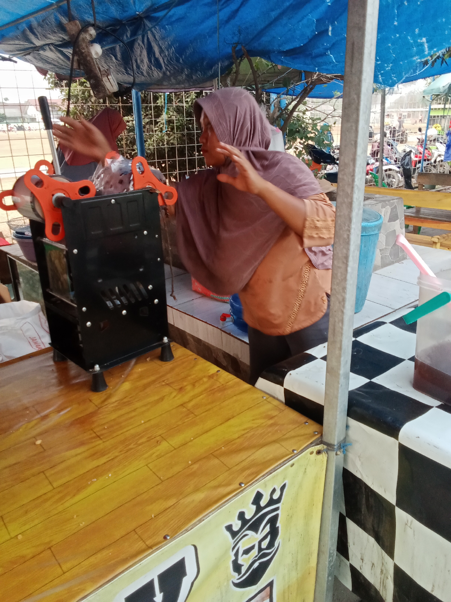 A woman wearing a purple hijab and an orange shirt stands behind a wooden counter. She is operating a black cup sealing machine with orange accents. The machine is on the left side of the frame. The counter is covered with a yellow wooden-patterned laminate. In the background, a blue tarp covers part of an outdoor stall, and trees and more stalls are visible. The scene appears to be an outdoor market.