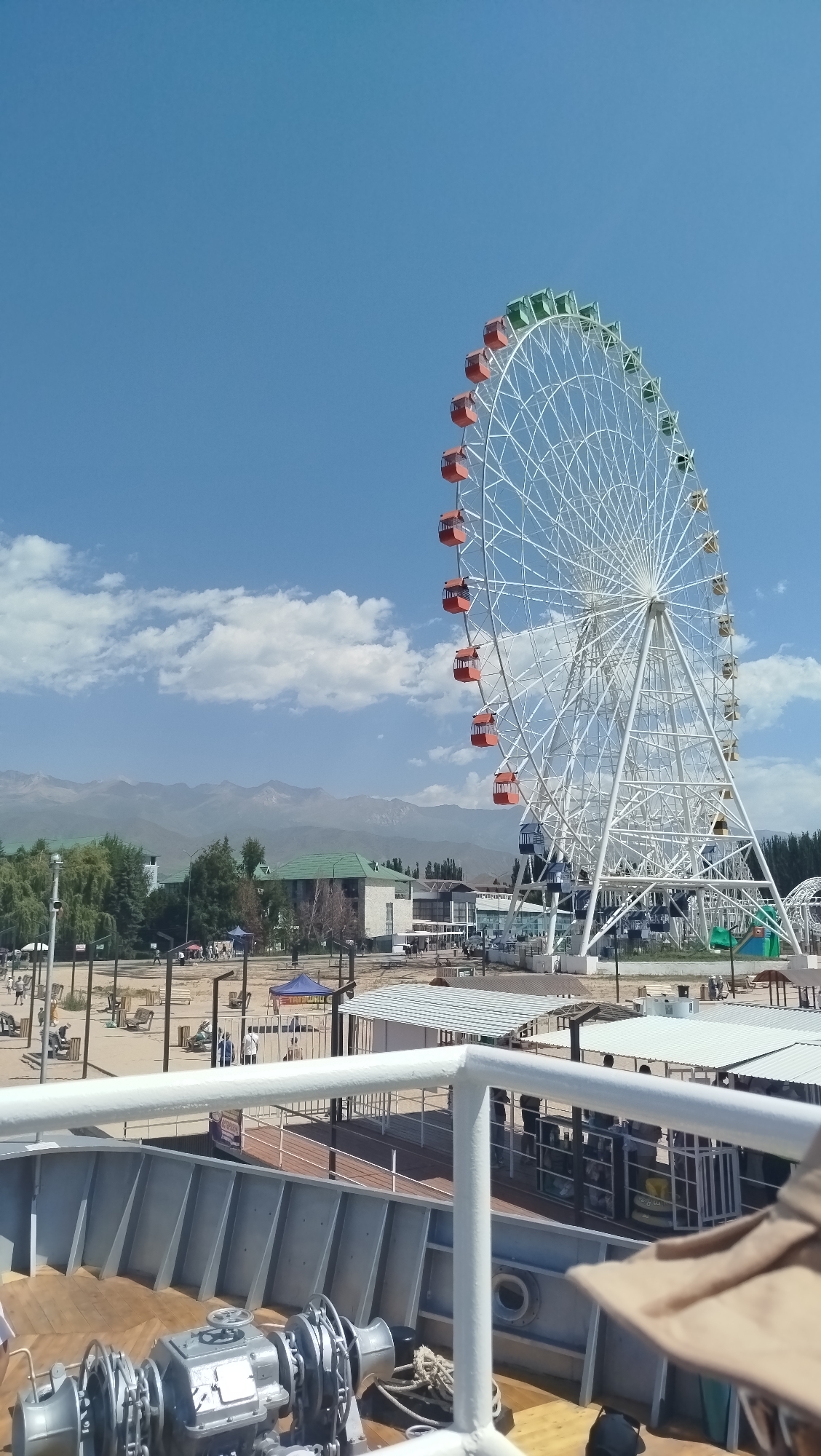 The image depicts a sunny day with a bright blue sky, featuring a large Ferris wheel as the dominant element. The Ferris wheel is white with colorful cabins, some red and some green, attached to its spokes. In the background, mountains rise against the horizon, suggesting a location near a range of hills or mountains. Several buildings with green roofs are visible, along with trees and a sandy area that might be a beach or park. In the foreground, part of a boat or ship is visible, including its deck, railing, and some machinery. There are a few people scattered around the area, some appearing to be walking or standing near the Ferris wheel and buildings, while others are seated on benches in the sandy area. The overall scene suggests an amusement park or recreational area on a clear day.