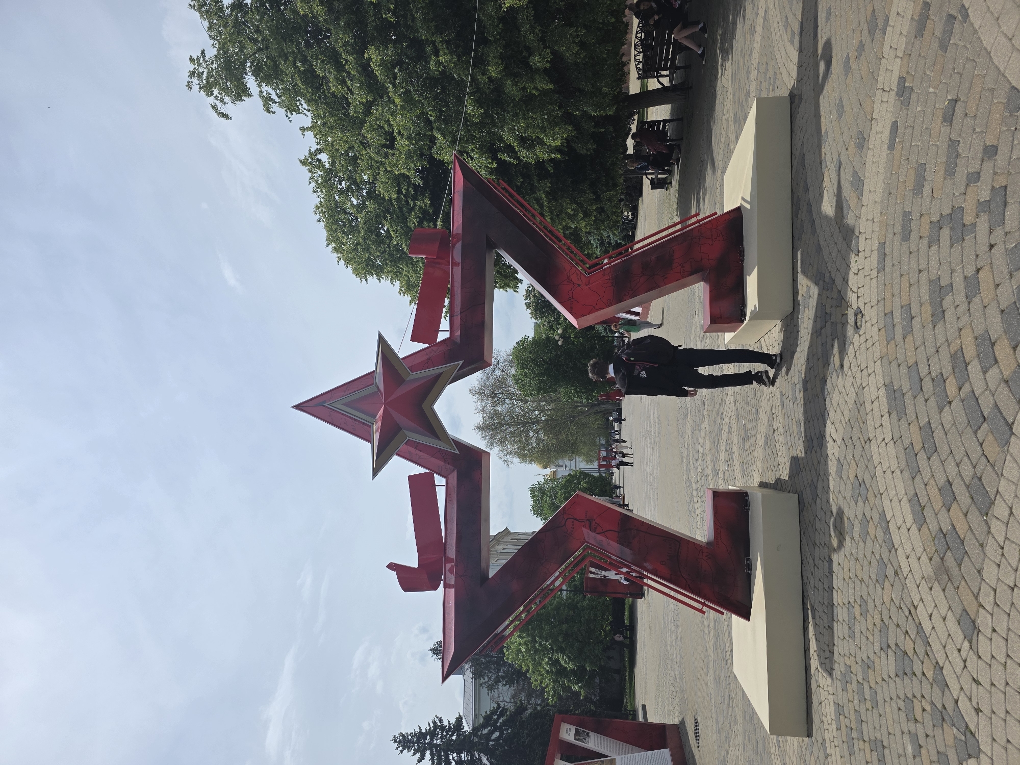 The image shows a large, red, five-pointed star sculpture with additional red geometric shapes attached, standing in a paved public square. The square appears to be in a park or pedestrian zone with trees and benches in the background. A person wearing a black jacket and pants walks near the star. Other people sit on benches further in the background. The pavers are multi-toned. This is a memorial in the city of Krasnodar, Russia.