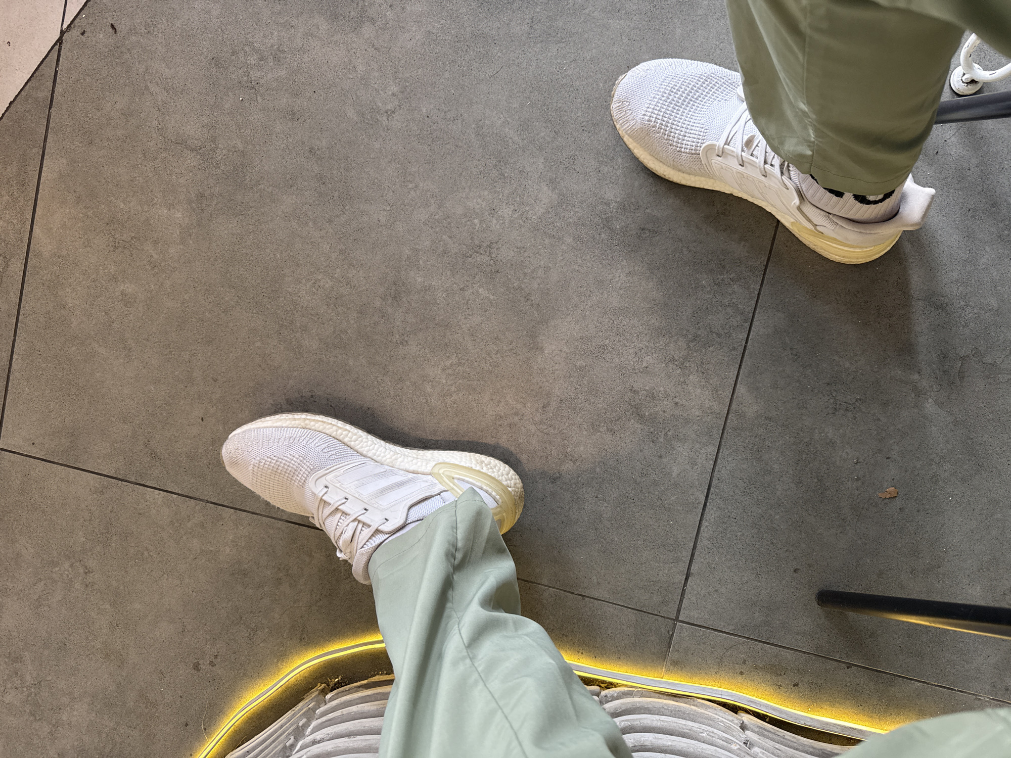 The image shows a person's view of their legs and shoes from a seated position. The person is wearing light green pants and white athletic shoes with visible white socks. The shoes have a distinct textured sole. The floor is grey tile with dark grout lines. The bottom edge of a chair is visible with a yellow neon light strip accenting its curve. This scene is unlikely to be identifiable as specifically within the city of Nha Trang, Vietnam, without further context.