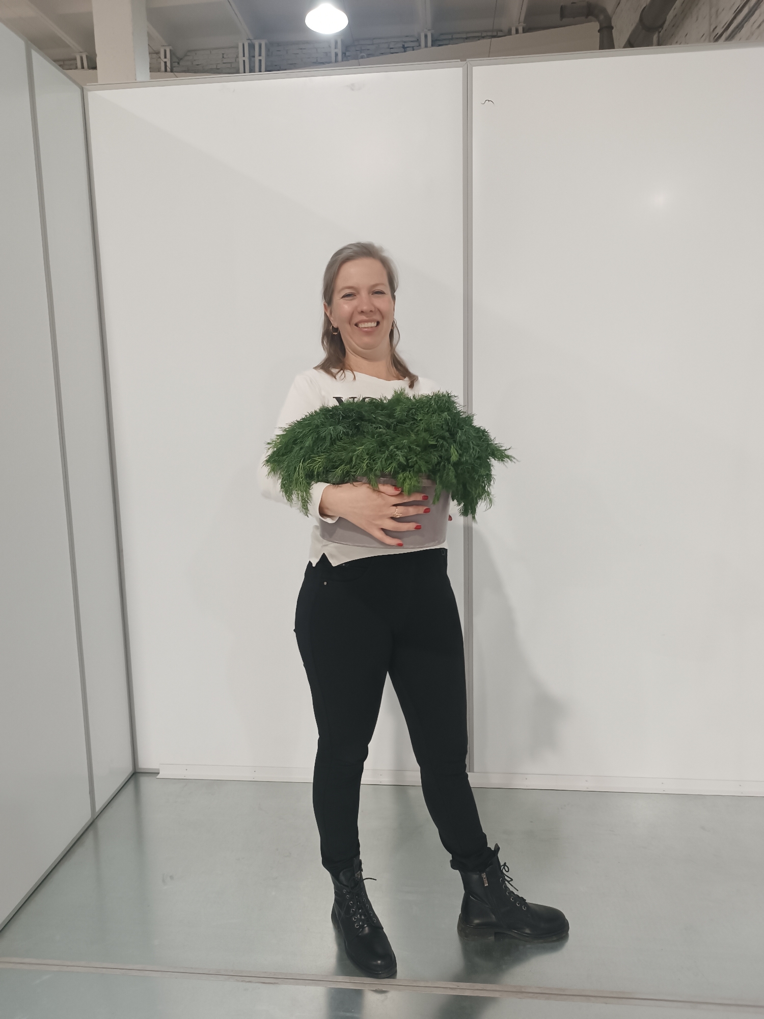 A woman with blonde hair is standing in an indoor location. She is holding a pot full of lush green plants. The woman is wearing a white long-sleeved shirt, black pants, and black lace-up boots. She is smiling at the camera.
The background consists of white walls divided by grey panels. The floor is a reflective grey material. An overhead light illuminates the scene.
There is no definitive evidence within the image to confirm the specific location as the city of Chaikovsky, Russia.