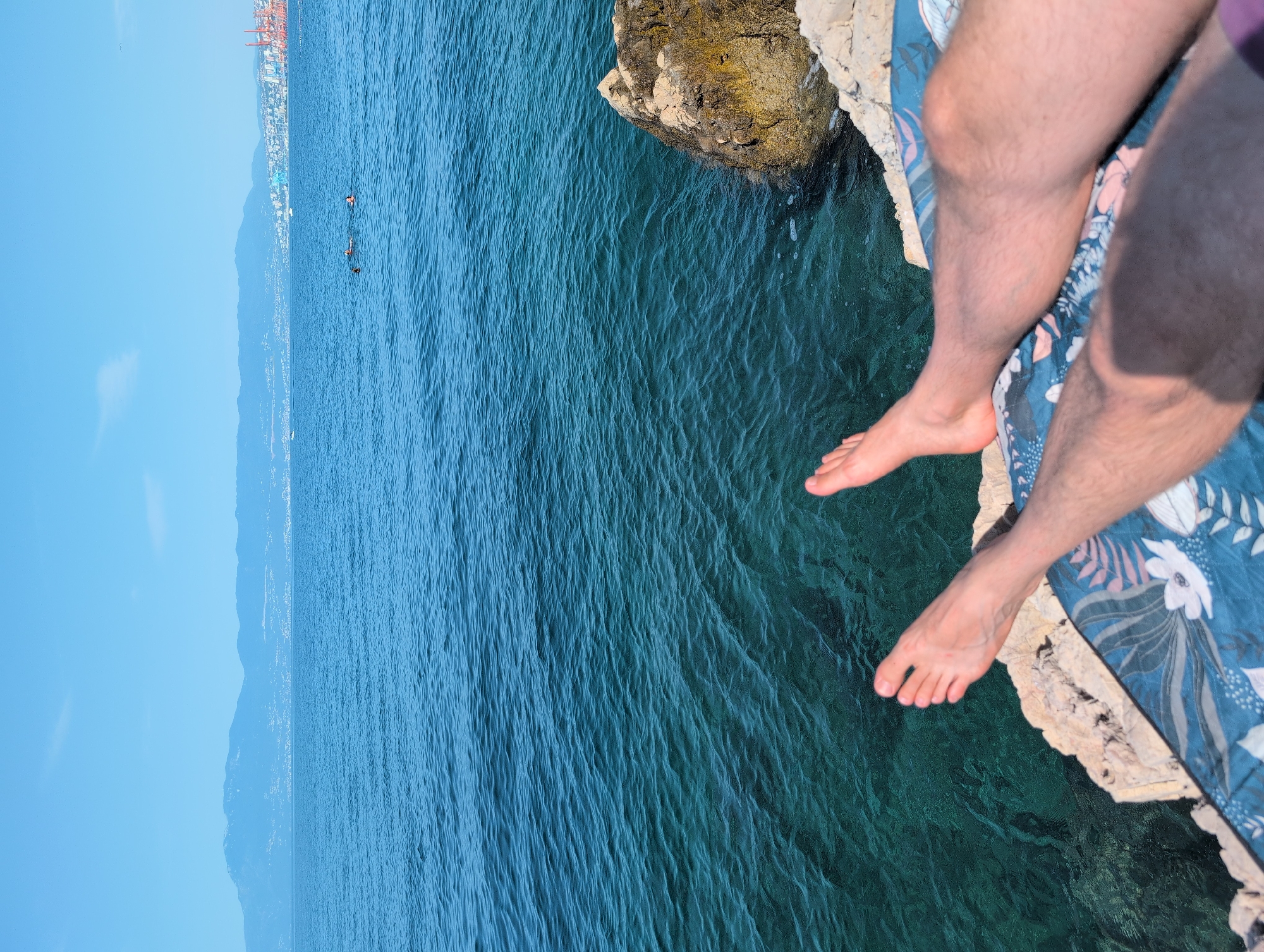 The image depicts a person's legs and feet resting on a patterned blanket at the edge of rocky terrain overlooking a clear, blue-green sea. In the distance, a coastline with a city and mountains under a bright blue sky is visible. Several people are swimming or wading in the sea further out. The scene is set during daylight hours, suggesting a warm climate. The location is identified as Kostrena, Croatia, a coastal town known for its scenic views and access to the Adriatic Sea.