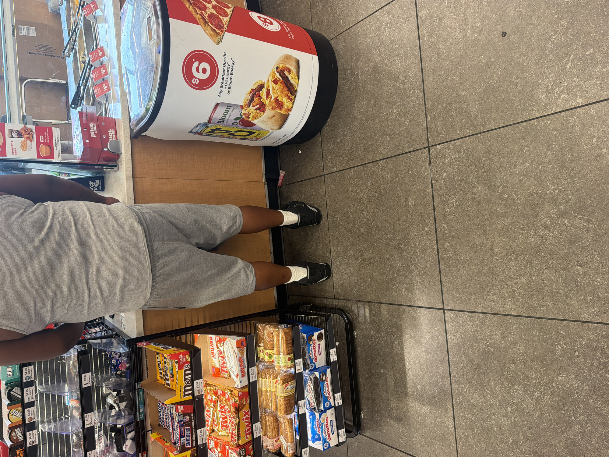 In Hayti, United States of America, a person stands in a convenience store. The individual is wearing a grey tank top and grey shorts, with white socks and black athletic shoes. They are positioned in front of a refrigerated display case containing various food items, and to the right of a cylindrical advertisement promoting breakfast burritos and energy drinks. In the background, shelves are stocked with candy bars, snacks, and other convenience store merchandise. The floor is tiled in a neutral tone.