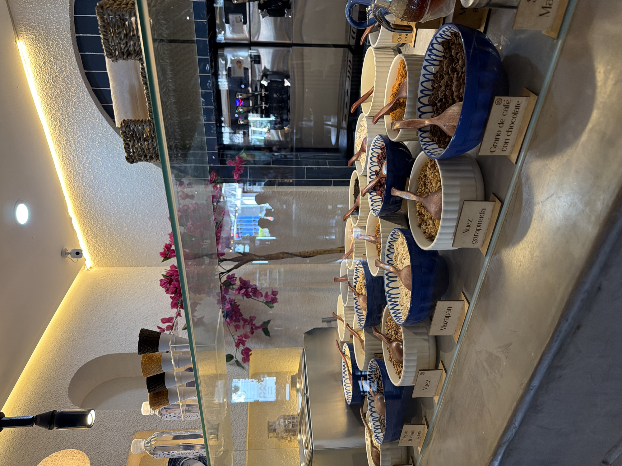 The image displays an indoor scene, likely within a cafe, dessert shop, or ice cream parlor, showcasing an array of toppings or ingredients behind a clear glass display case. The setting features white textured walls, warm artificial lighting, and blue tiled accents visible in the background reflections.
A series of white and blue-patterned ceramic bowls are arranged on a counter, each filled with a different topping and containing a small wooden scoop. Visible labels for these items include "Chispas chocolate OSCURO" (Dark chocolate chips), "Nuez" (Nut/Walnut), "Mazapán" (Marzipan), "Nuez garapinada" (Candied nut), and "Grano de café con chocolate" (Coffee bean with chocolate). Other bowls contain various grains, nuts, or cereals. Partially visible text reads "Qui cor" and "Ma".
Reflections on the glass display show elements of a service area or kitchen, including what appears to be a coffee machine or similar equipment. A security camera is mounted high on a textured wall. Decorative elements include a woven basket on a shelf, a branch with pink artificial flowers, and several plastic water bottles alongside stacks of ice cream cones (black and brown varieties) stored below the main display. The scene suggests a self-service or assisted-service station for customizing food items.