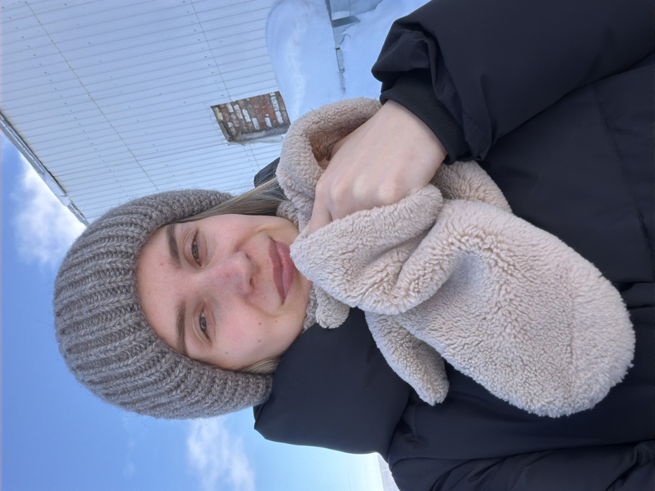 A woman wearing a gray knit cap and a black puffer jacket poses outside in winter. She holds a soft, light beige scarf or muff near her face. Her skin appears fair. The sky is visible, partially obscured by a white rooftop and a chimney. The photo is taken at an unusual angle.