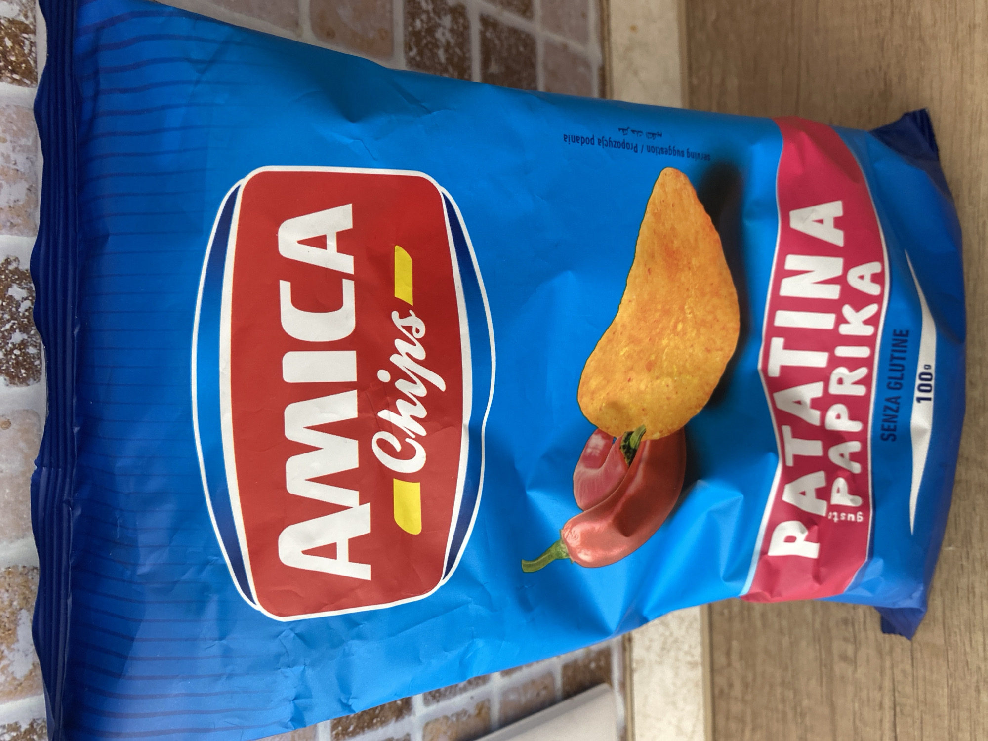A bright blue bag of "Amica Chips" flavored with paprika is the primary subject of the image. The bag is positioned on a wooden surface, likely a table or counter, with a tiled backsplash visible in the upper portion of the frame. Two red chili peppers and a single potato chip are depicted on the front of the bag, suggesting the flavor profile. Text on the bag includes "AMICA Chips," "PATATINA PAPRIKA," "GUSTO," "SENZA GLUTINE," and "100g." There are also some Arabic characters and the words "serving suggestion / Propozycja podania" vertically oriented on the right side of the bag. The lighting is natural and appears to be from the side, casting a slight shadow. There are no people visible in the image. The scene is indoors and appears to be a still life setup for product photography. The image does not provide any information to suggest it is located in Chișinău, Moldova, or any other specific city.