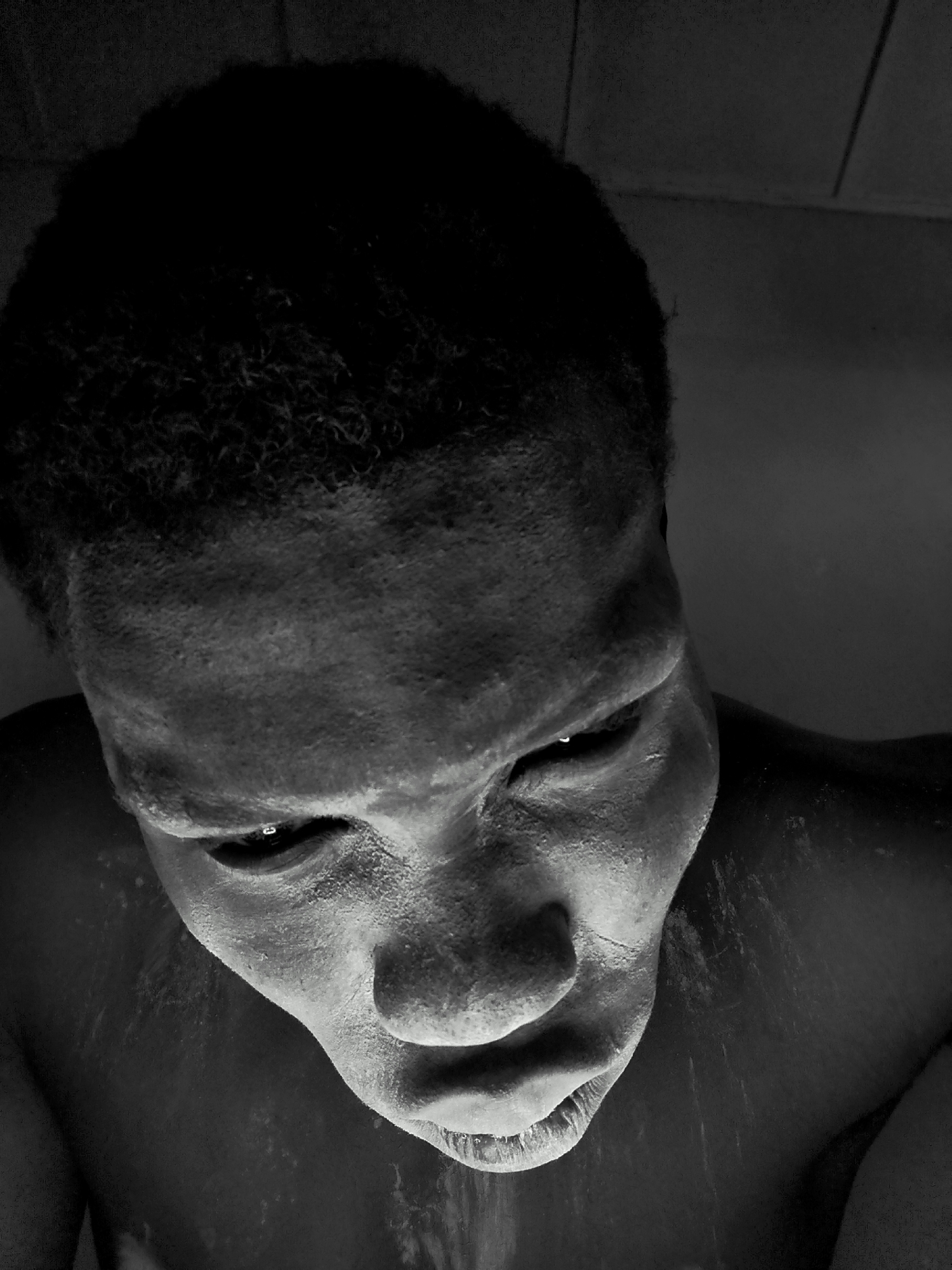 The image is a high-angle, black and white close-up of a person's face, powdered with a white substance. The person is looking downwards, with their eyes partially closed. The skin appears textured and slightly damp in places. The person has dark, curly hair. The background is dark and out of focus, suggesting an indoor, dimly lit environment. There are no discernible landmarks or environmental features that would allow for the specific location of Sabon Tasha, Nigeria to be identified from the image alone. The image focuses solely on the subject's face and upper torso.
