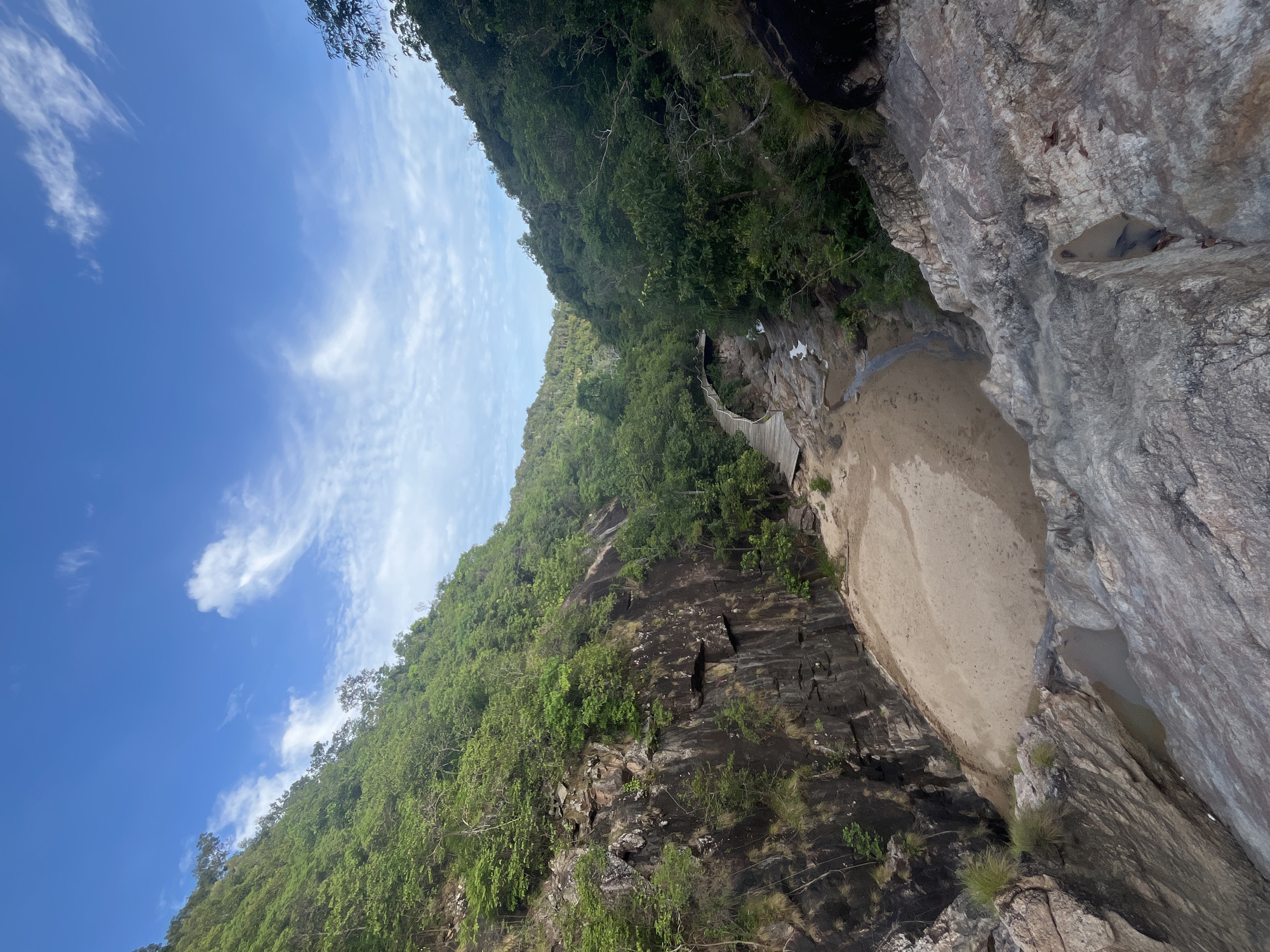 The image captures a natural scene in Koh Phangan, Thailand, looking down into a rocky gorge. A small stream flows through the gorge, with a sandy bank visible on one side. A wooden boardwalk curves along the side of the gorge, leading into the dense green foliage that covers the steep slopes. The sky above is a vibrant blue, dotted with wispy white clouds. The foreground is dominated by large, textured rocks, suggesting the perspective is from a higher vantage point, possibly a cliff or overlook. The overall environment is lush and verdant, indicative of a tropical setting. There are no people or animals visible in the image.