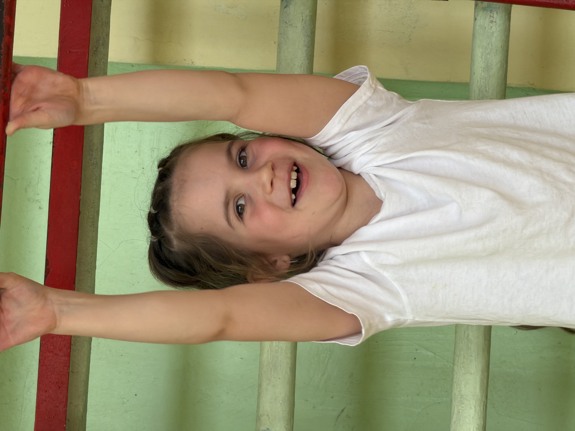 A girl in a white t-shirt hangs upside down from a ladder with red and green rungs, smiling at the camera. The background is a green wall with some yellow patches. The location is Izhevsk, Russia.
