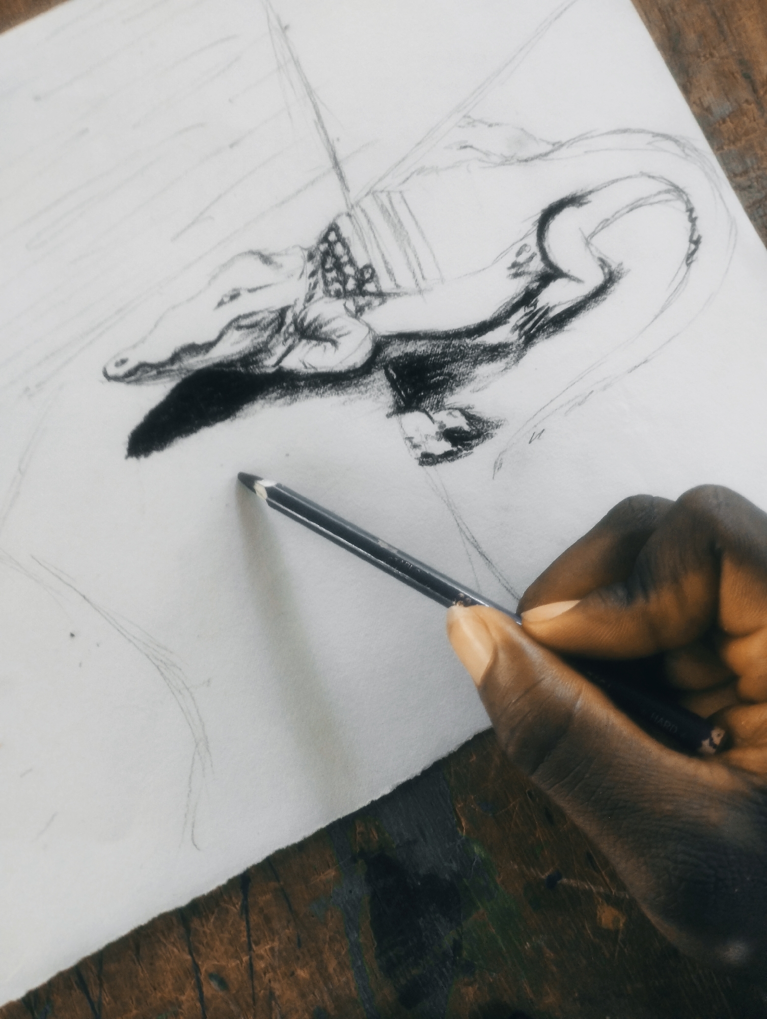 A close-up, overhead shot shows a hand holding a pencil and sketching on a piece of paper. The sketch depicts a dragon-like creature with wings and a long tail, with its head facing left. The creature is partially shaded with charcoal, and it appears to be wearing a saddle. The paper is on a wooden surface that appears to be a table or desk. The background is blurred, but suggests an indoor setting.