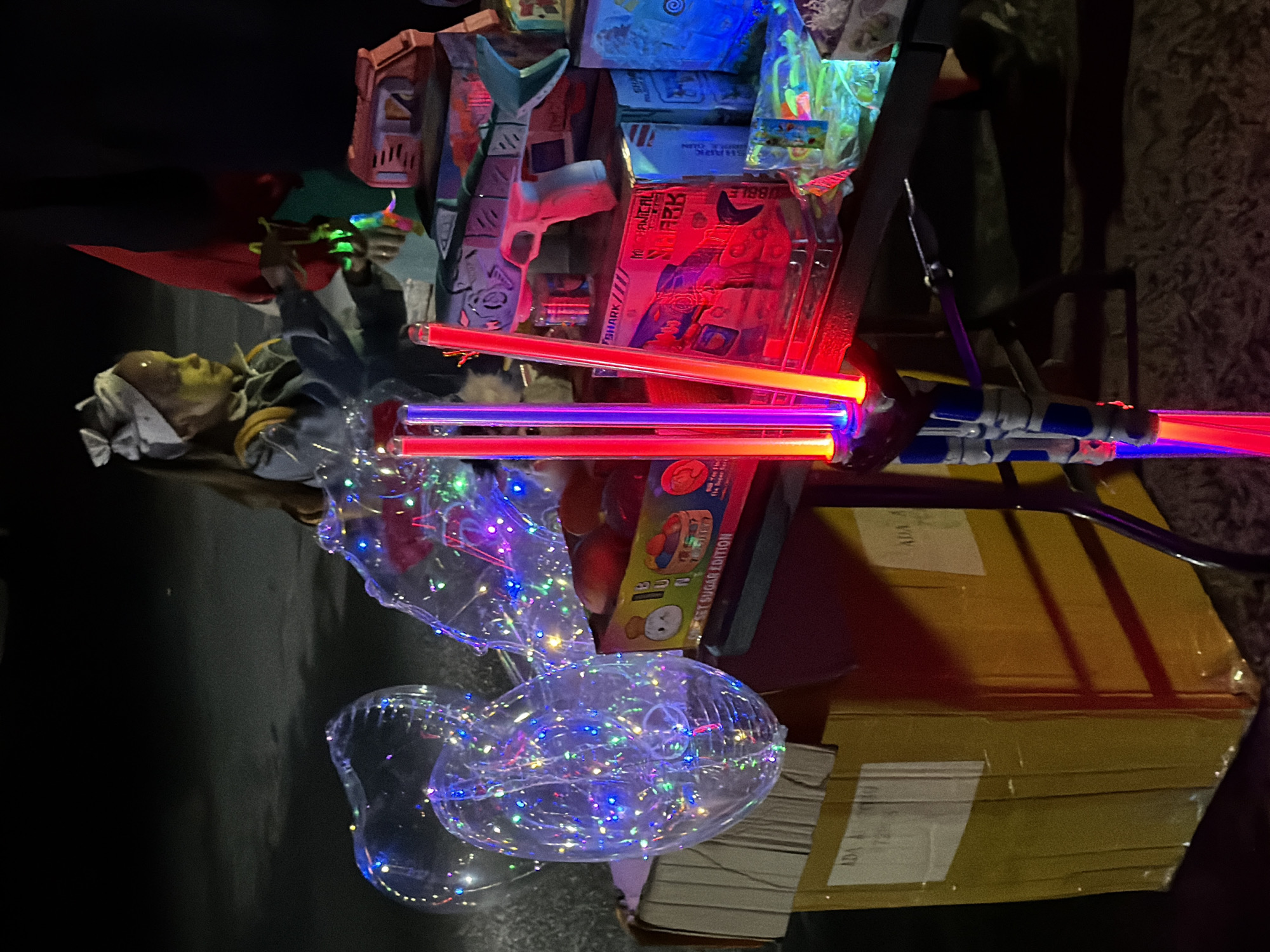 In Unknown, Unknown, a young girl with light-colored hair, wearing a light blue outfit and a white bow in her hair, is visible in the background. She has yellow face paint and is holding a green toy. In the foreground, there are several objects, including large, clear balloons filled with colorful LED lights, a yellow cardboard box, and a table displaying various toys. Among the toys are two light-up wands emitting red and yellow light, a toy shark gun that is light blue and gray with red accents, and other packaged toys with colorful designs, some of which are illuminated. The scene appears to be outdoors at night, with a dark background.