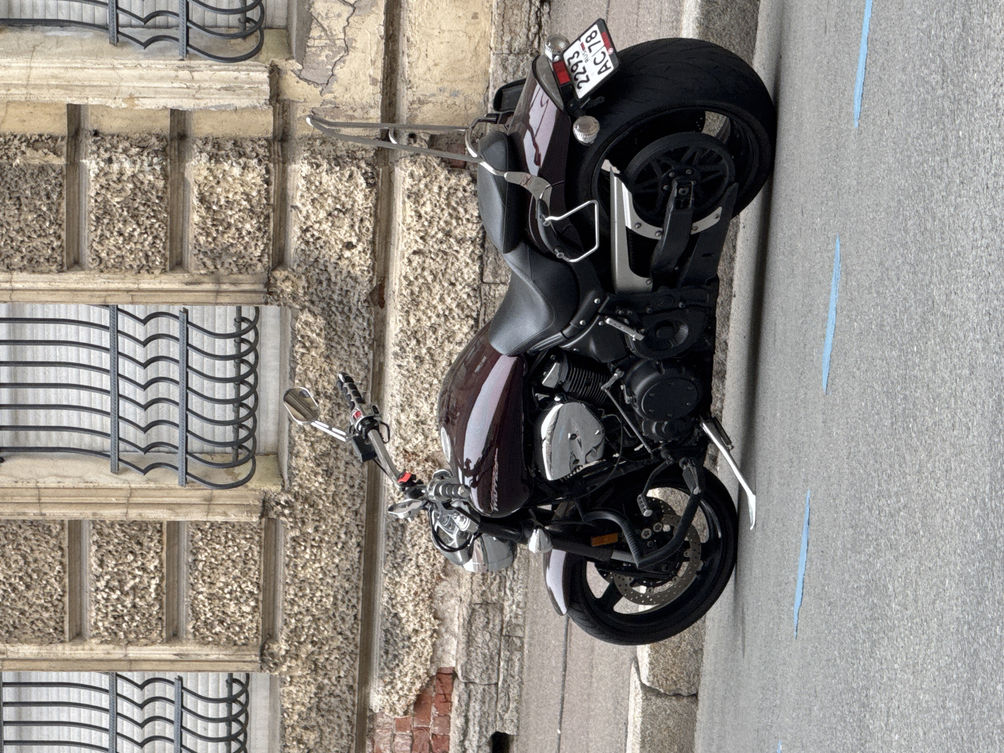 The image depicts a dark maroon motorcycle parked on a gray asphalt street, near a building with ornate stonework and wrought iron window grills. The motorcycle faces the camera, with its front wheel slightly turned towards the right. A Russian license plate is visible on the back of the motorcycle. The location is likely Saint Petersburg, Russia, given the license plate and architectural style. The scene is static, with no evident actions or interactions. The environment is urban and appears to be a typical street scene.