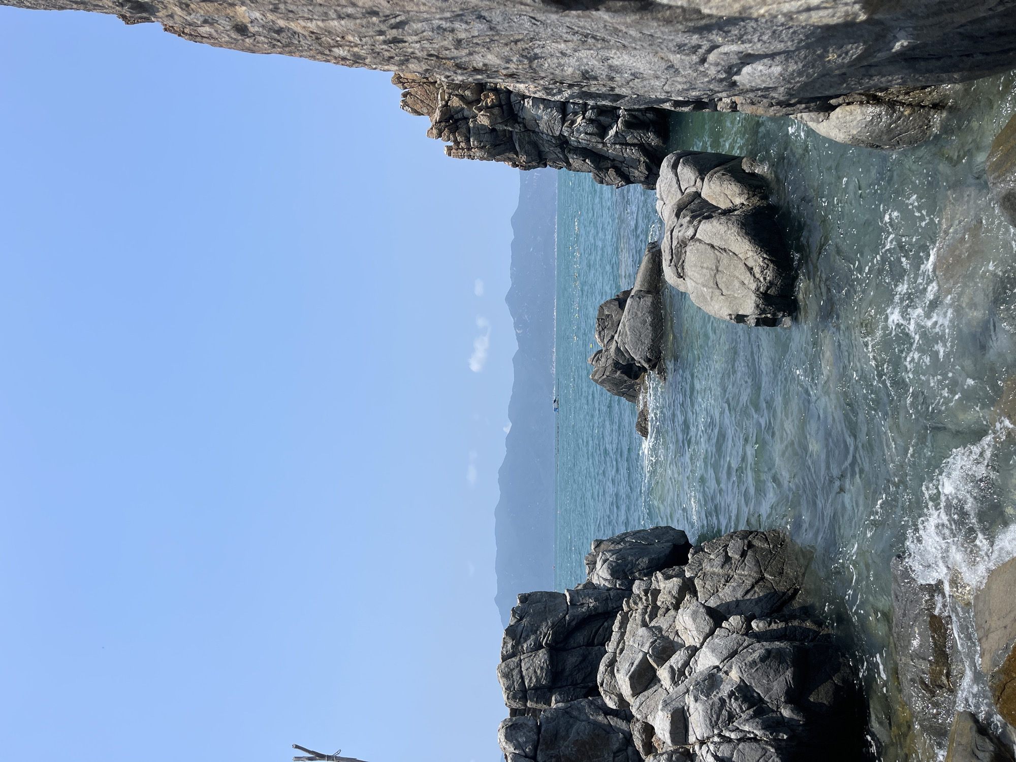 The image shows a view of the sea from a rocky cove in Vĩnh Lương, Vietnam. Large, gray rocks frame the scene on the top and bottom, with the clear, turquoise water of the sea filling the space between them. The water is slightly choppy, with some white foam visible near the rocks. In the distance, a faint mountain range can be seen across the water under a clear blue sky.