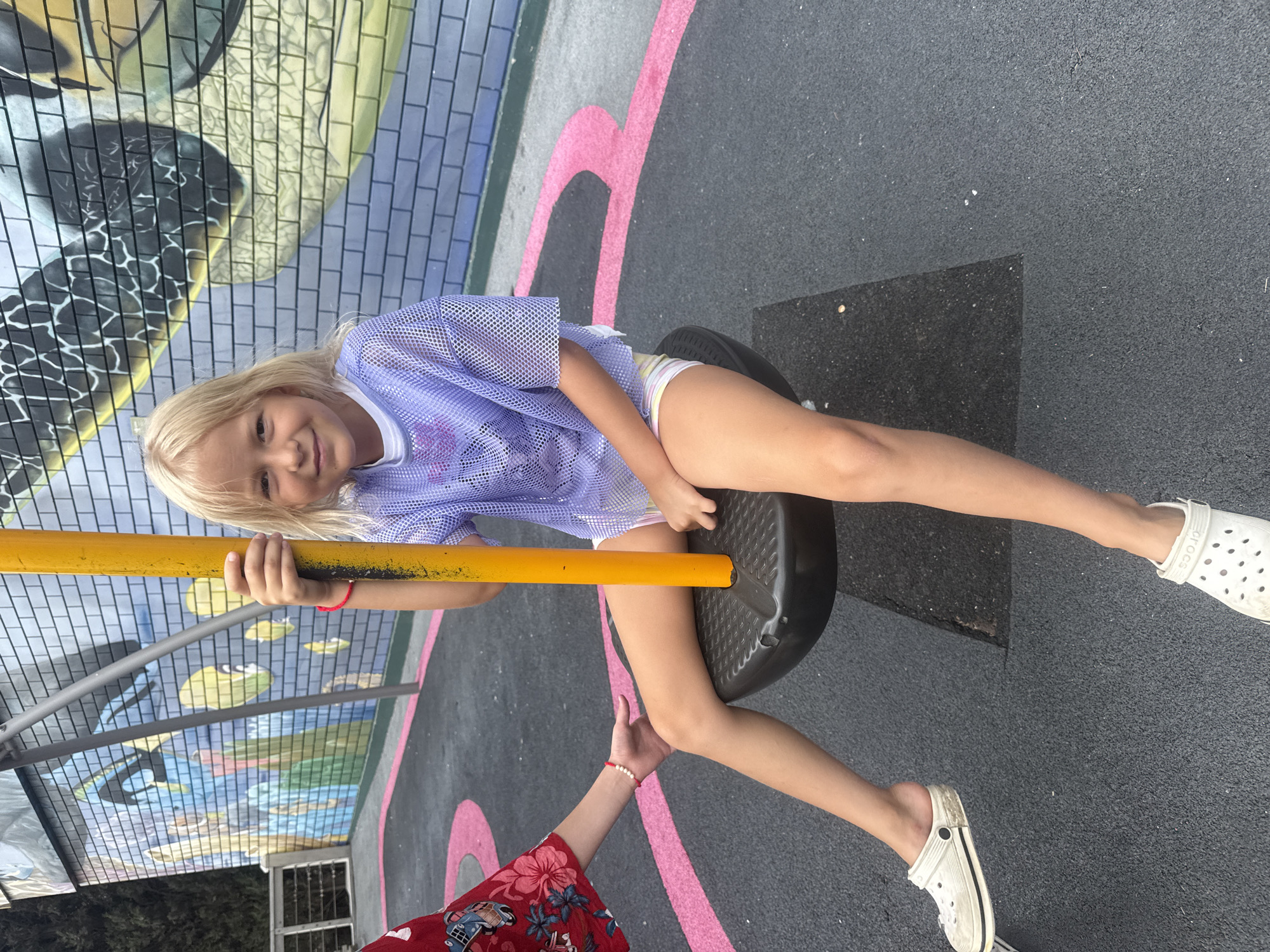 A young girl with blonde hair and a purple mesh shirt is sitting on a playground swing. She is wearing white Crocs and colorful striped shorts. Her legs are crossed at the knees, and her feet are resting on the ground. Her right hand is resting on the yellow pole of the swing, and her left hand is partially visible on the right side of the frame. Another person's arm, wearing a red shirt with a floral pattern, is visible on the left side of the frame, reaching towards the girl. The playground has a black rubberized surface with pink markings. In the background, a brick wall is painted with a colorful mural. The image appears to be taken outdoors during the day.