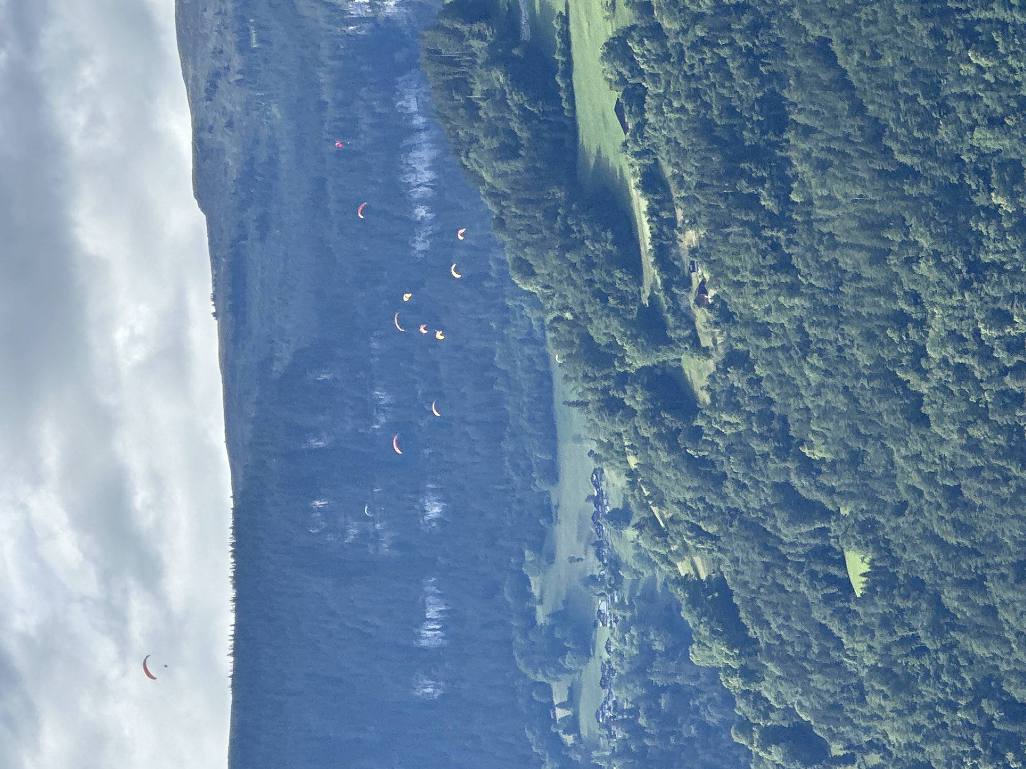 This is a vertical, eye-level, aerial photograph taken from a high vantage point. The image captures a mountain slope and a forest, with several paragliders in the air. The paragliders, predominantly in red and white colors, are scattered across the sky, appearing as small dots against the vast landscape. The scene is set against a backdrop of a rugged mountain on the left, with patches of white cloud in the sky, and a dense, green forest on the right. A small, bright green field is visible within the forest. The lighting suggests it is daytime, likely afternoon, with some clouds in the sky. The overall mood is one of adventure and freedom, as the paragliders soar through the open air. No text is visible in the image. The location is Unterseen, Switzerland.