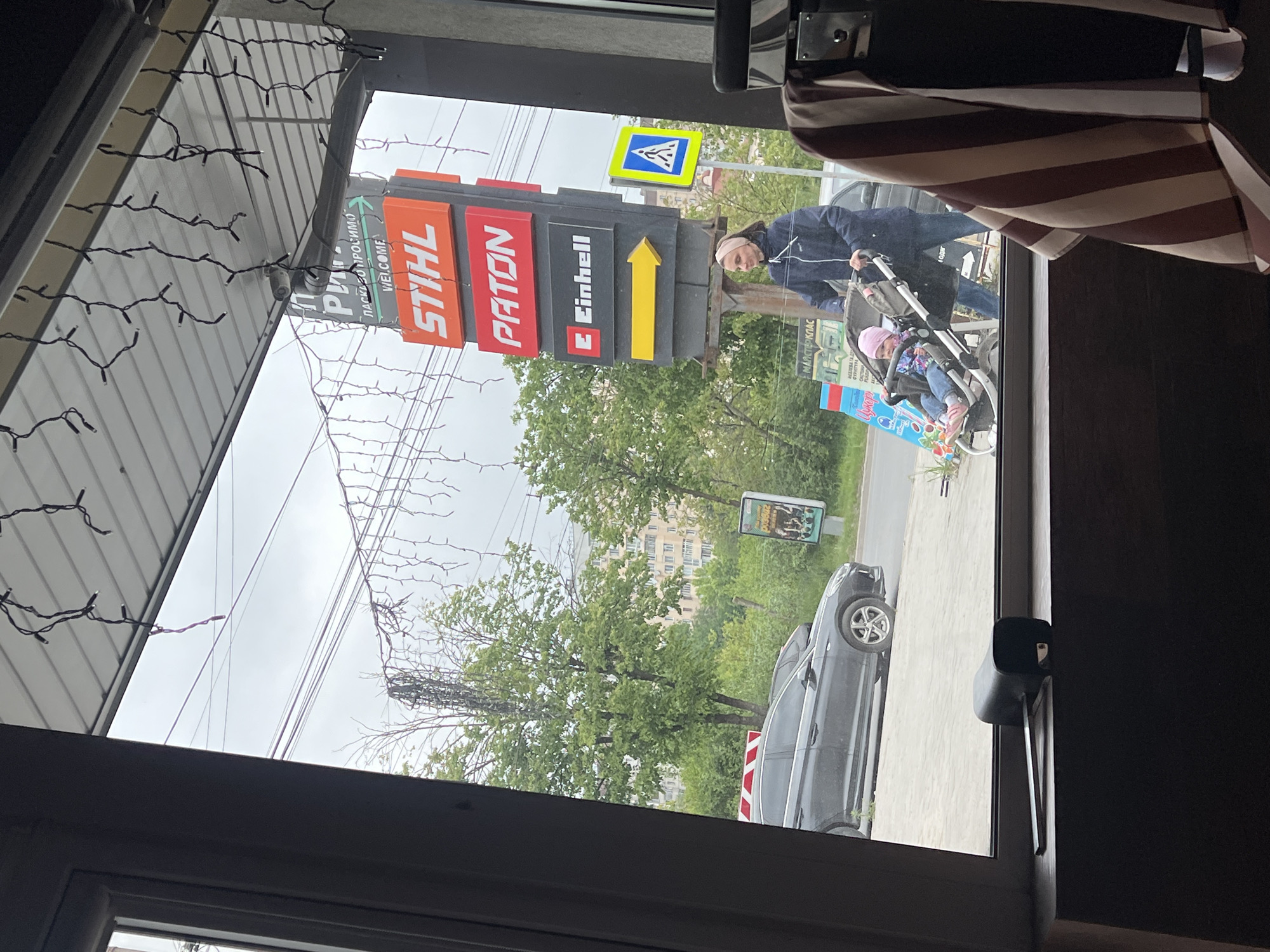 The image, shot from inside a building through a window, shows a street scene in Ternopil, Ukraine. Prominently visible through the glass is a tall, vertical sign displaying the logos of Stihl, Paton, and Einhell, each associated with a yellow arrow. A pedestrian crossing sign can also be seen.
An elderly woman, wearing a dark jacket, is pushing a stroller with a child in it on the sidewalk. The street is populated with trees and a gray vehicle parked on the side of the road. A distant building can be seen past the trees.
The window itself is framed by the dark interior of the building. String lights are affixed on the building’s exterior.