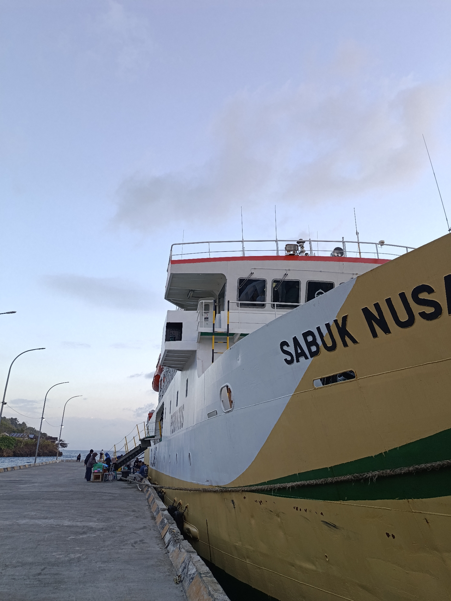 The image captures a close-up perspective of a large ferry docked at a pier. The ferry, painted in white and gold with a green stripe, has the name "SABUK NUS" clearly visible on its hull. A thick rope secures the vessel to the pier. In the background, a few people are gathered on the pier, some appearing to be waiting to board or disembark. The pier itself is made of concrete, with lampposts visible. To the left, a rocky, vegetated coastline meets the water. The sky above is a pale blue with scattered clouds, suggesting it might be early morning or late afternoon. The location is identified as the city of Mansalean, Indonesia.