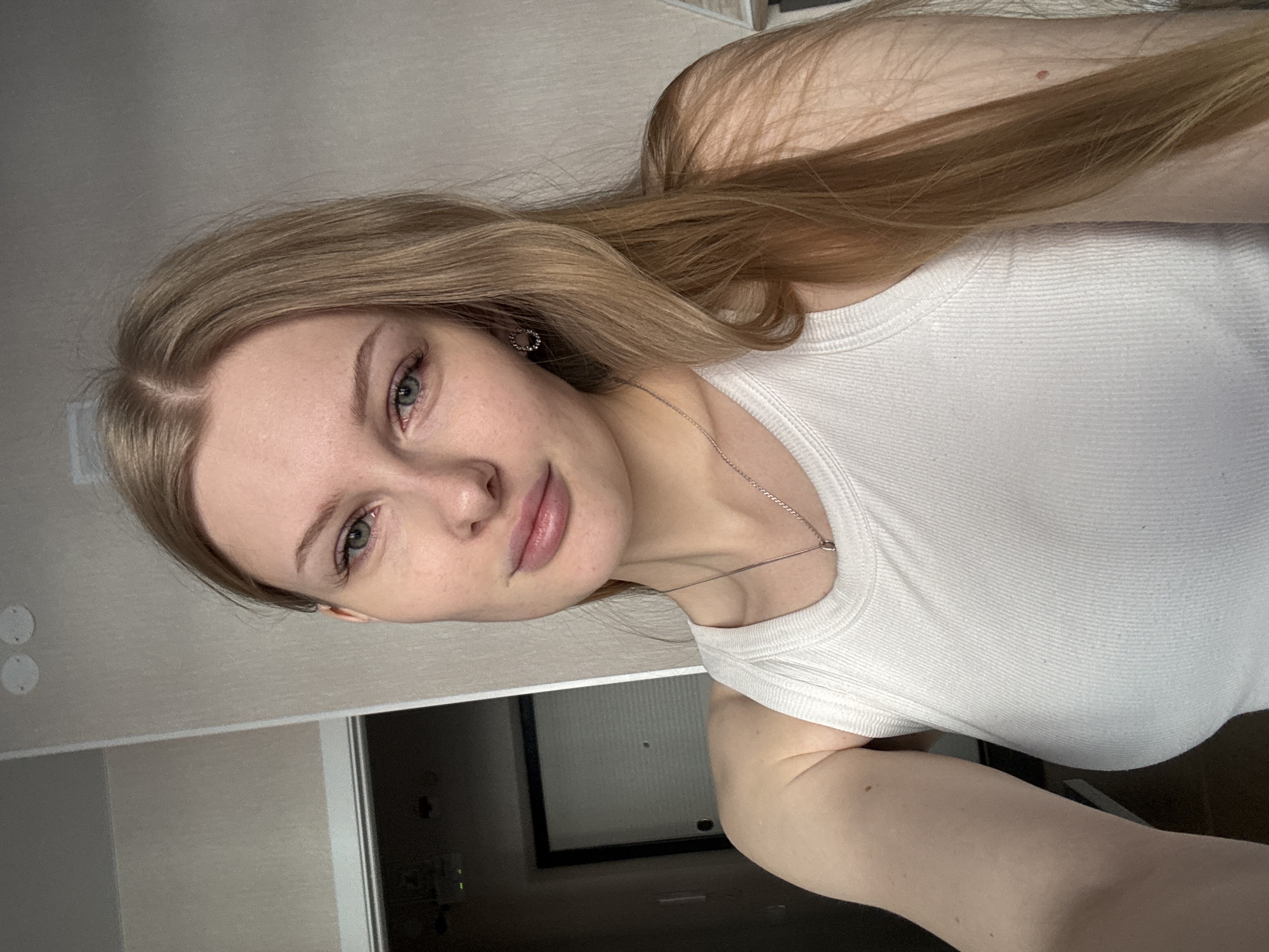 A fair-skinned young woman with light brown, long hair and blue-green eyes poses indoors. She wears a white tank top and a thin silver necklace. The background shows a white wall, a doorframe, and what appears to be a sliding closet door with a black frame. The location appears to be a residential interior, although the precise location within the city of Perm, Russia cannot be determined from the image alone.