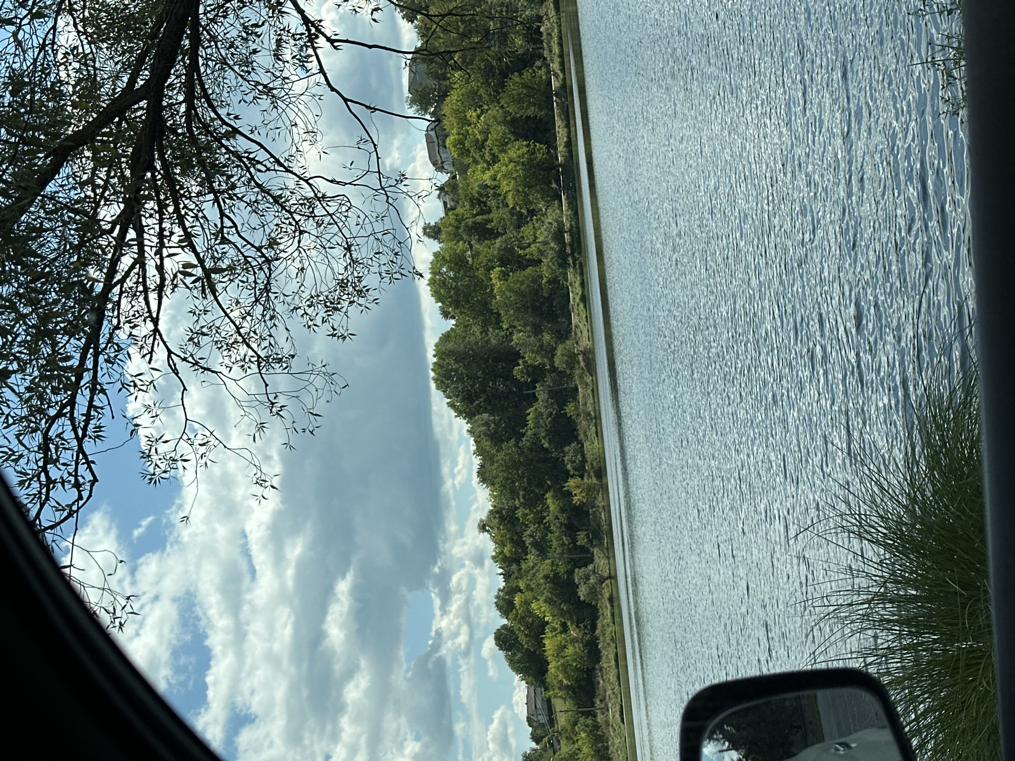 The image captures a tranquil lake scene in Makiv, Ukraine, viewed from the interior of a vehicle. In the foreground, the side mirror of the vehicle is visible on the left, reflecting the sky and trees. Lush green grass grows along the edge of the water. The lake itself is a prominent feature, its surface rippled by a gentle breeze, reflecting the blue sky and scattered white clouds.
Beyond the lake, a dense line of trees forms a verdant backdrop, with scattered houses nestled amongst the foliage on a gentle slope. The trees display a variety of green hues, indicating a healthy, mature forest. The sky above is a vibrant blue, punctuated by large, fluffy white clouds that cast soft shadows on the landscape. A large tree branch with delicate leaves extends into the frame from the top right, partially obscuring the sky and adding a natural framing element to the composition. The overall impression is one of serene natural beauty and a peaceful rural setting.