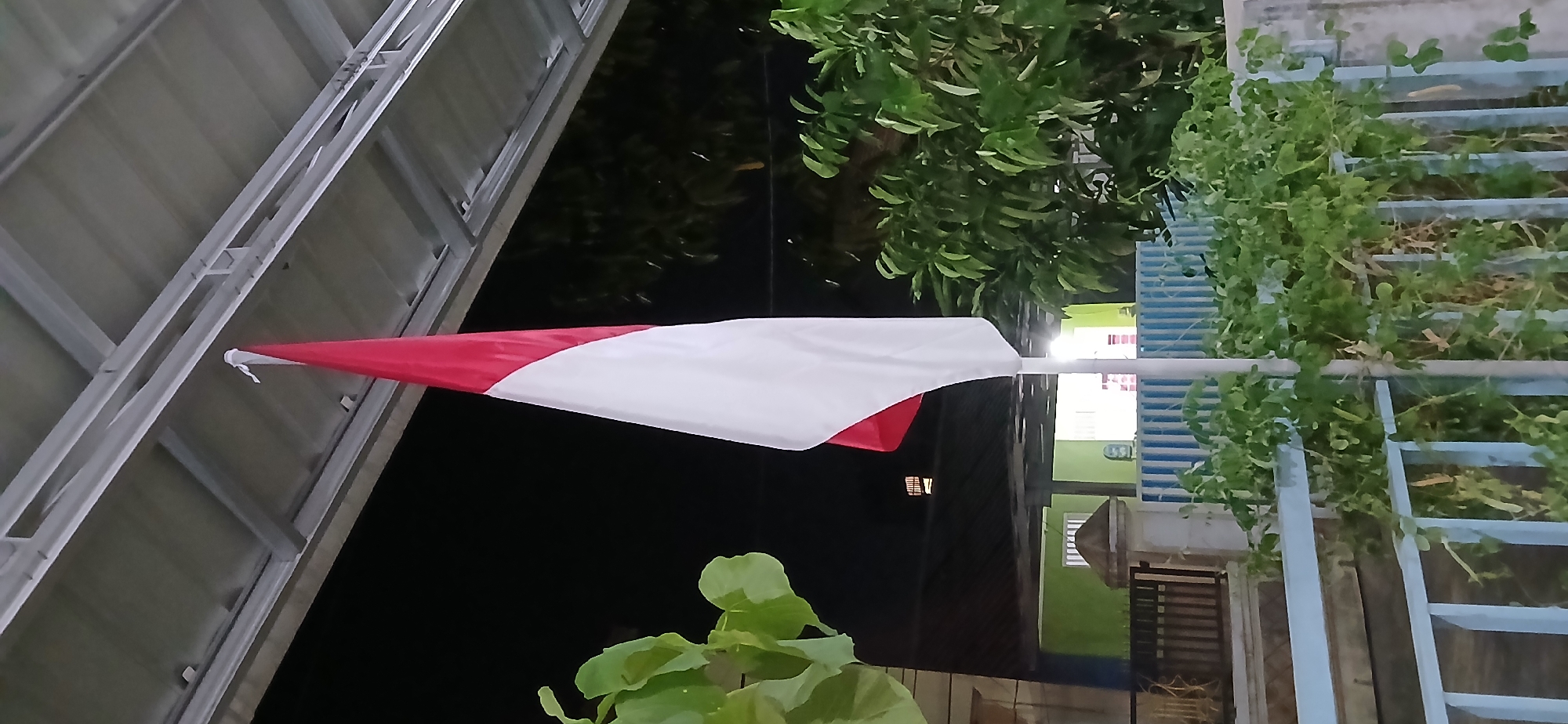 The image shows an Indonesian flag, red and white, flying from a flagpole. The flag is positioned centrally in the frame, partially obscured by foliage. Behind the flag, a building with a green exterior and windows is visible, suggesting a residential or institutional setting. The scene is outdoors at night, indicated by the dark sky and artificial lighting from the building. The flag is attached to a white pole which extends upwards, disappearing beyond the top edge of the frame, where it is secured to a metal structure, likely a roof or awning. In the foreground, a light blue metal fence and green plants are present, with some of the plants trailing over the fence. The overall impression is of a flag flying at a private residence or a local establishment in Samarinda, Indonesia, possibly during an evening ceremony or a display of national pride. The location is a city in Indonesia, as suggested by the national flag.