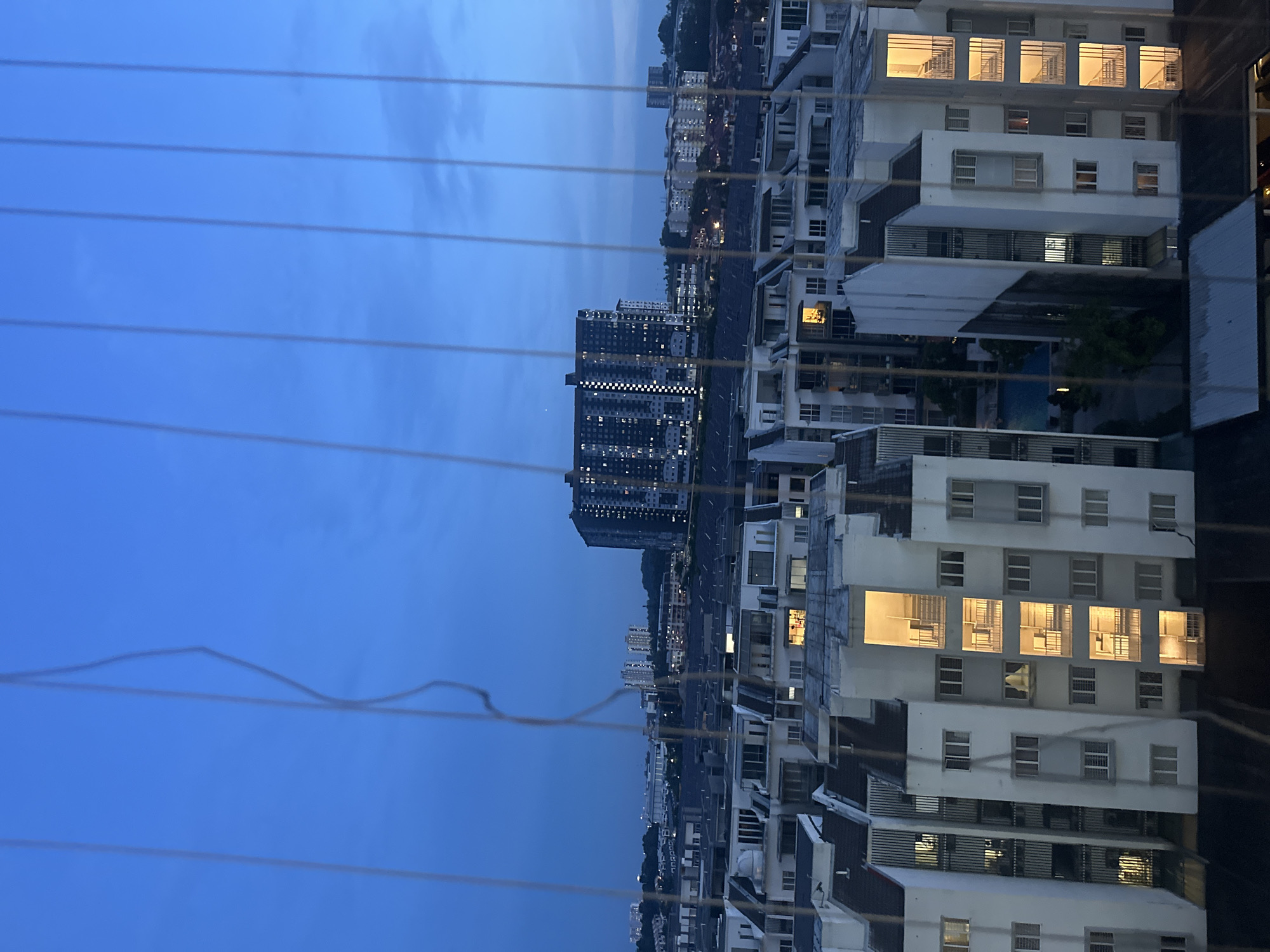 The image presents an elevated view of Subang Jaya City Council, Malaysia, at dusk. A series of utility lines are visible in the foreground. The cityscape is composed of residential buildings, primarily white in color, with illuminated windows. A high-rise apartment building stands prominently in the center of the view. Streetlights and car lights indicate active roadways. The sky is a gradient of blue, suggesting the time of day. No people are visible in the frame.
