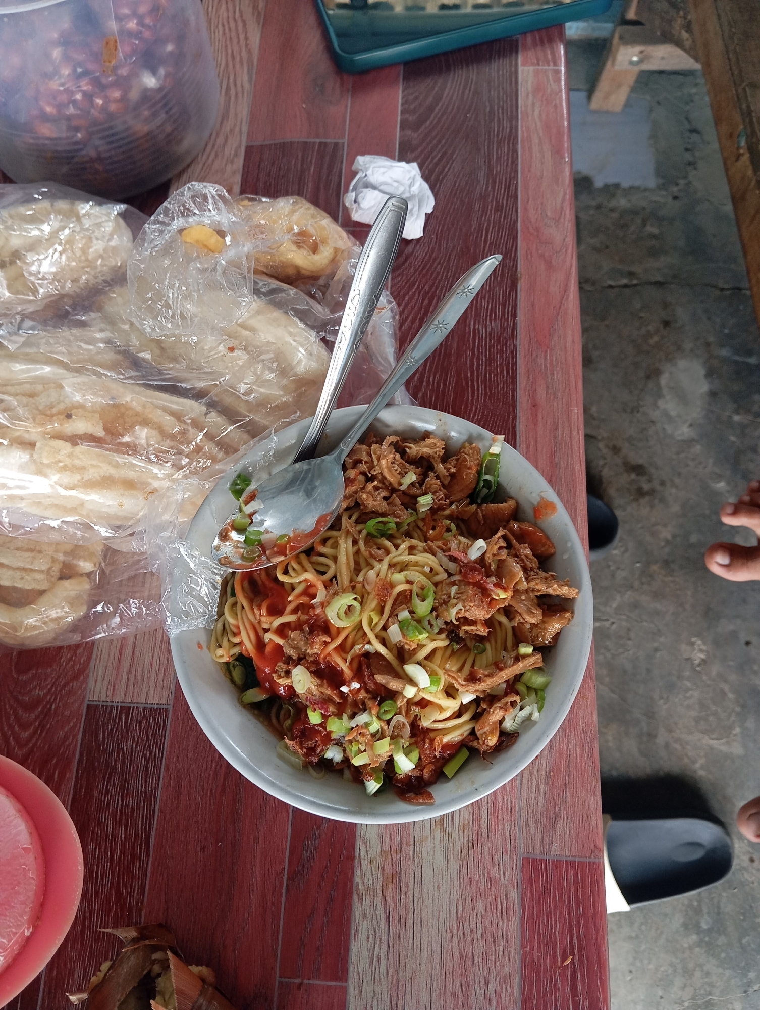 In a dimly lit eatery in Sokaraja Wetan, Indonesia, a solitary bowl of noodles sits on a weathered, reddish-brown wooden table. The noodles, a mix of fine strands, are topped with shredded chicken, finely chopped green onions, and a generous dollop of red chili sauce. Two spoons, one silver and one with a patterned handle, rest in the bowl, suggesting that the meal is either being prepared for consumption or has been partially eaten.
To the left of the noodles, a stack of light brown, crispy crackers, possibly krupuk, are sealed in a clear plastic bag. Further back and to the left, a large, transparent plastic container overflows with what appears to be dried peanuts. The surrounding environment is utilitarian, with a concrete floor visible in the background, a partially seen black sandal, and a sliver of a wooden bench. A pair of feet, one bare and the other clad in a dark sandal, are visible in the lower right corner, implying the presence of at least one person out of frame. The scene evokes a sense of humble sustenance, a common sight in local Indonesian eateries.