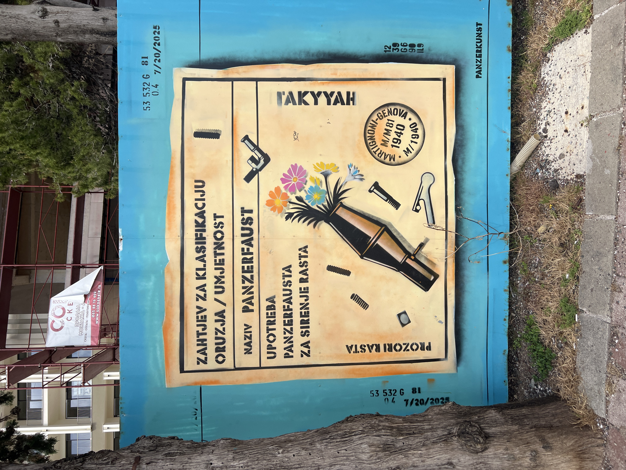 A blue shipping container stands on the street in Rafailovići, Montenegro. The container is adorned with a painted mural of a faux shipping document, which details the classification of the artwork as a "Panzerkunst" piece. The document outlines the classification of arms/art, with a description of the object as a Panzerfaust intended for spreading Rasta. The document is also decorated with flowers growing from the Panzerfaust and the text "TAKYYAH". The painted document also features faux inspection stamps from "Martignoni-Genova M/M81 1940 M/1940". Serial numbers and other container markings are painted on the container. A tree trunk is visible at the bottom, and a construction scaffolding with a banner advertising scaffolding sales is visible on the left.