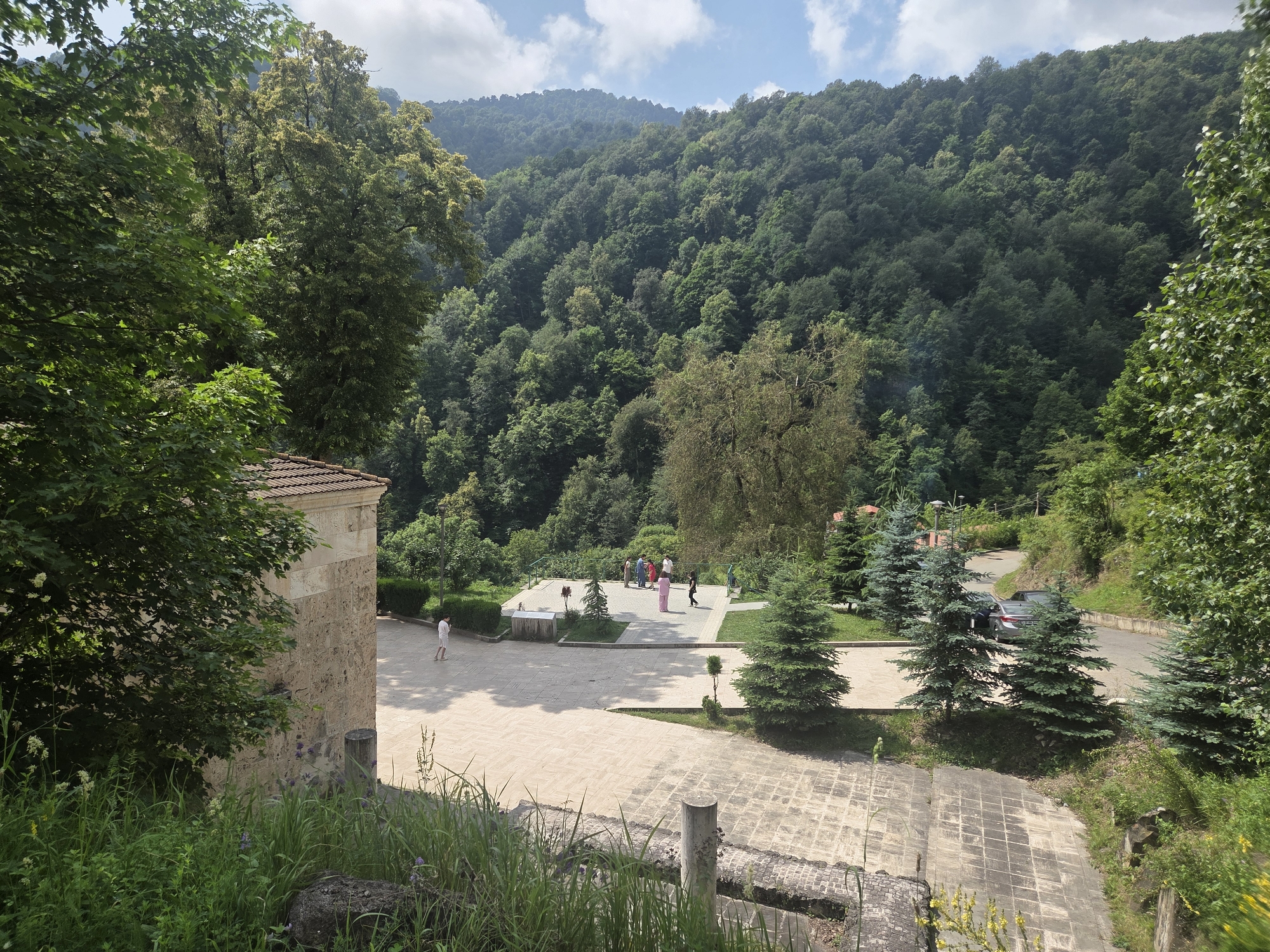 This is a high-angle, wide shot of an outdoor public space surrounded by lush, green, mountainous terrain. In the foreground, to the left, is a portion of a building with a terracotta tiled roof and light-colored stone walls. In the middle ground, a paved area with light-colored square tiles is visible, featuring several individuals standing and walking. Some of the individuals are dressed in casual attire, with a group of about five people gathered together. There are also several evergreen trees scattered across the paved area and the surrounding grass. To the right, a paved road winds its way up the hillside, with a silver car parked near some of the evergreen trees. The background is dominated by a dense forest covering the undulating slopes of the mountains, with a partly cloudy sky above. The scene suggests a serene and natural setting, possibly a park or a historical site, located in the city of Teghut, Armenia, given the context.