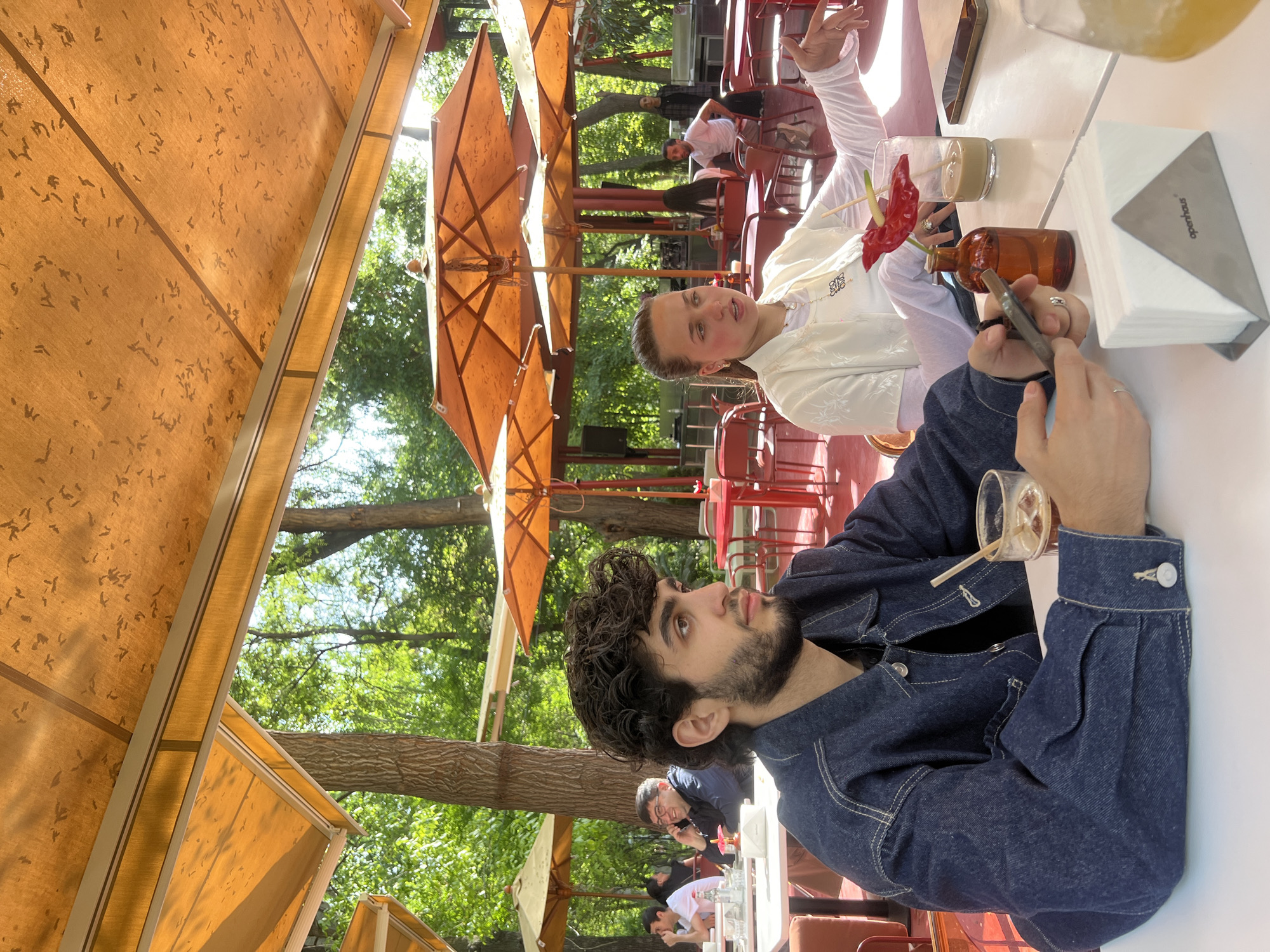 A man with dark curly hair and a beard is seated at an outdoor table. He is wearing a dark denim jacket and has a partially consumed beverage in front of him. Across from him is a woman in a white top, and she is holding her hand up. The table is cluttered with drinks, including a glass with a red flower garnish, a brown bottle, and a container of napkins. The restaurant is located outdoors, as indicated by the trees and umbrellas in the background. Other patrons can be seen in the background.