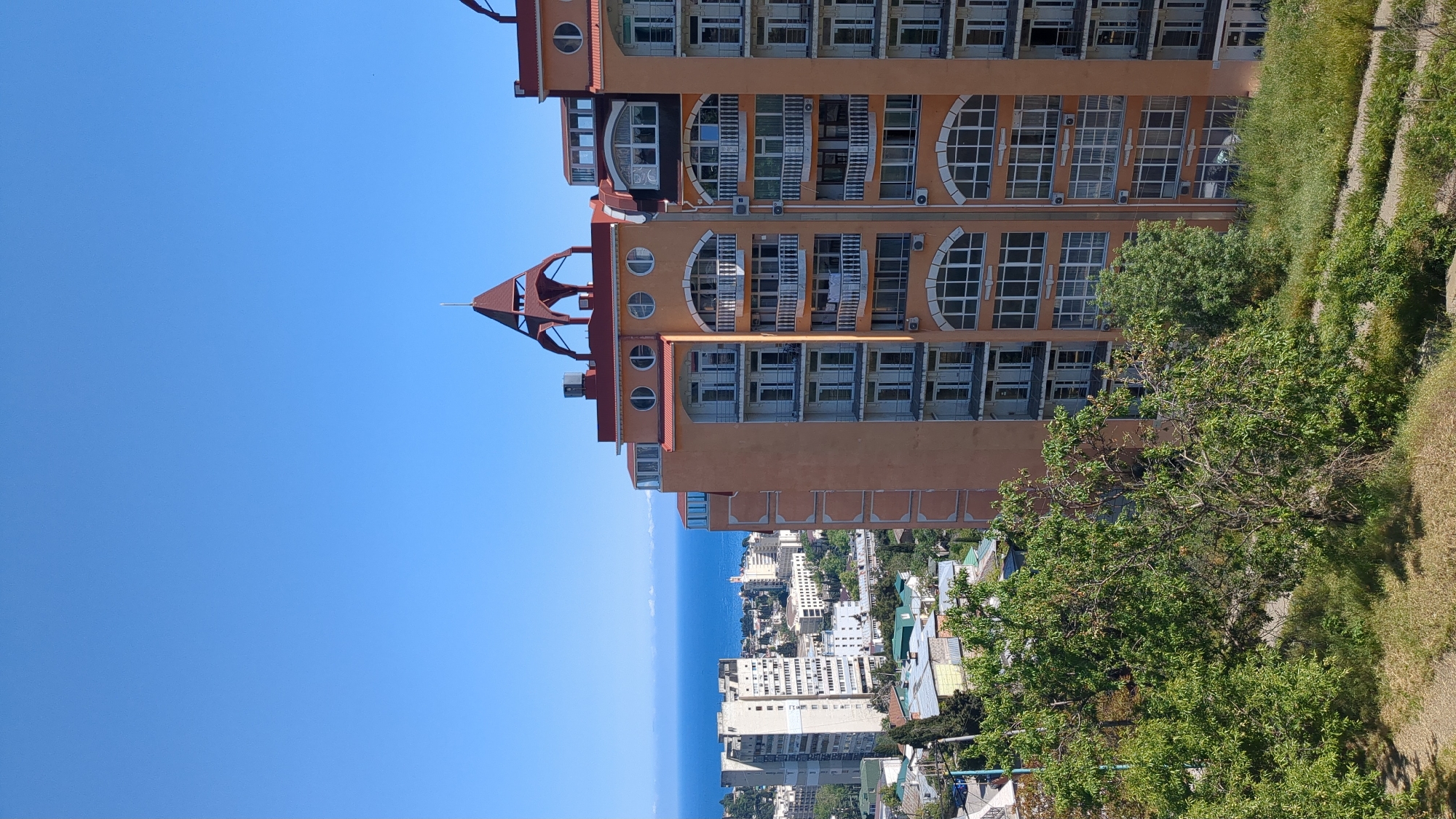 The image depicts a tall, peach-colored building with a red roof and ornate architectural details, including arched windows and balconies. It is located in город Ялта, Ukraine, as suggested by the surrounding urban landscape with other buildings visible in the distance. A body of water, likely the Black Sea, is visible in the background, partially obscured by the buildings. In the foreground, there is greenery, including trees and grass, suggesting a hillside or elevated vantage point from which the photo was taken. The sky is clear and blue, indicating a sunny day. There are no people visible in the image. The building appears to be residential or commercial, given the presence of balconies and windows.