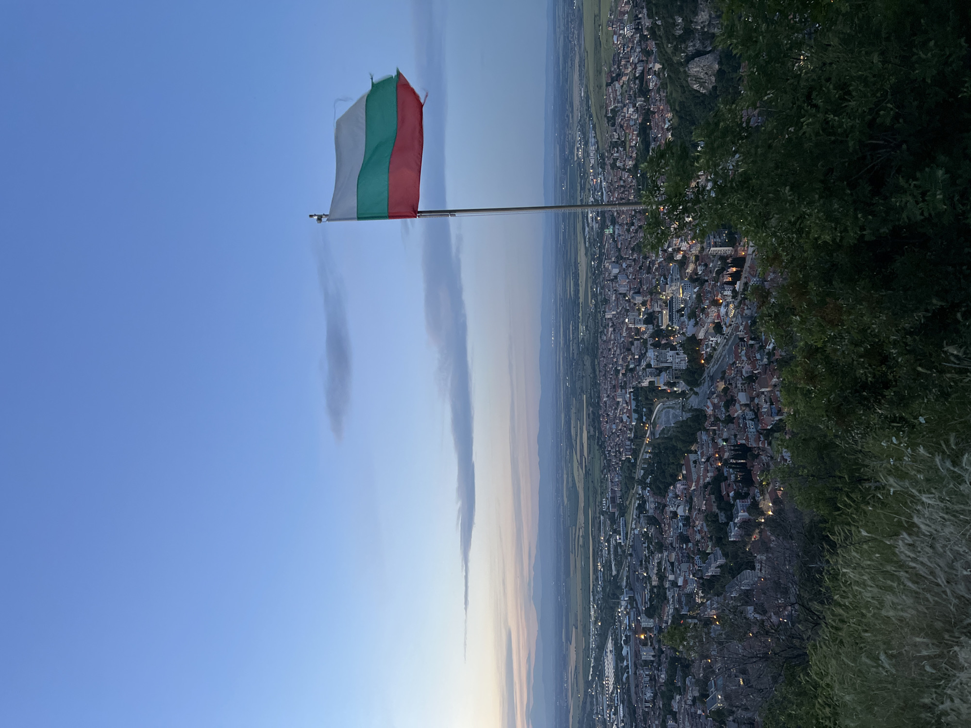 The image captures a view of Asenovgrad, Bulgaria, featuring the Bulgarian flag prominently displayed. The flag, consisting of white, green, and red horizontal stripes, is mounted on a pole positioned above the city. The city sprawls in the background, dotted with numerous buildings and illuminated by scattered lights, indicating it might be dusk or dawn. The foreground includes some greenery, possibly trees or bushes, adding depth to the scene. The sky is a gradient of blue, suggesting the time of day. Two vertical streaks in the sky, possibly clouds or contrails, add to the atmospheric perspective.